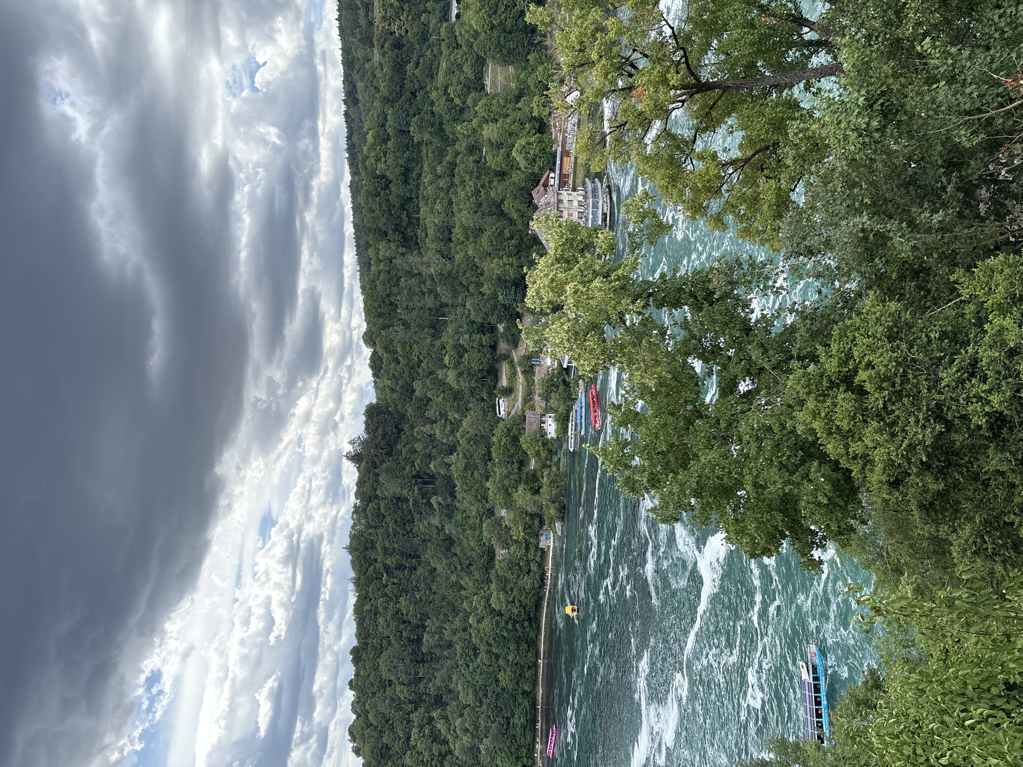 The image depicts the Rhine Falls in Dachsen, Switzerland, under a cloudy sky. The falls are characterized by turbulent, turquoise water with white foam, indicating strong currents. Several boats are visible on the water, including a bright pink boat on the left, a yellow boat in the middle, a blue and white boat, and a red boat on the right. A larger building, possibly a restaurant or visitor center, is situated on the riverbank. The surrounding landscape is densely covered with lush green trees, forming a backdrop to the falls. The sky is filled with dramatic, dark grey storm clouds, interspersed with patches of lighter clouds and glimpses of blue sky.