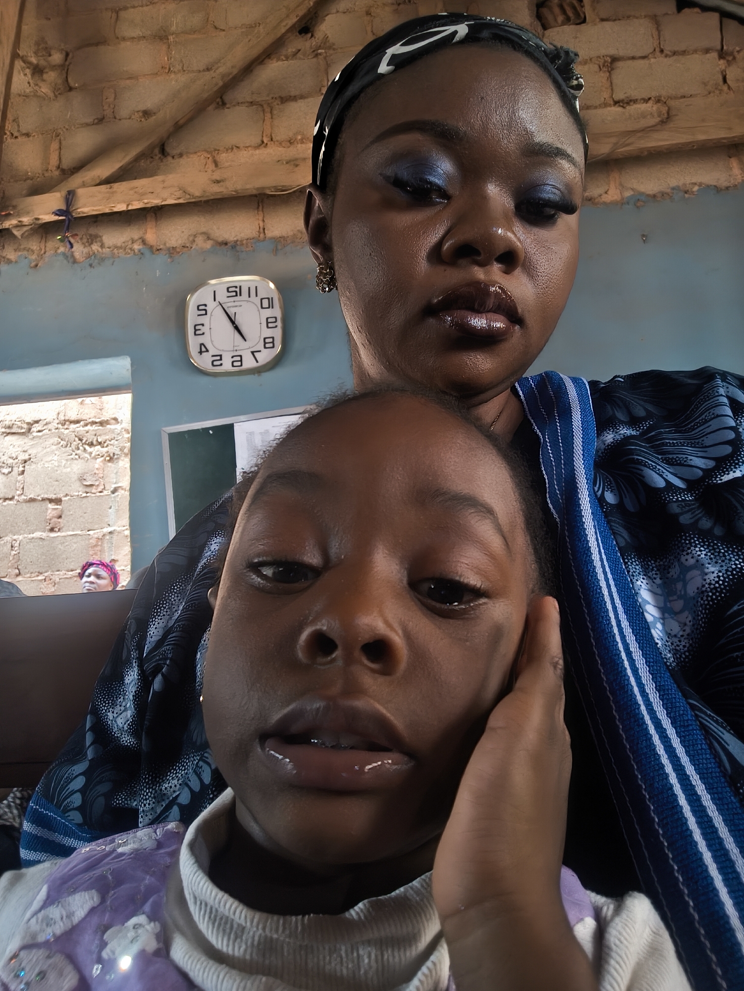 A medium-shot indoor portrait shows a woman and a young girl. The woman is behind the girl, slightly out of focus. She wears a dark headscarf with white patterns, blue eye shadow, and dark, glossy lipstick. A blue and white patterned shawl is draped over her shoulders. The girl in the foreground is holding her face with her right hand. She wears a white shirt with a light purple top. Behind them, the blue wall of the room has a square clock and a framed board mounted on it. The clock is white with black numbers and hands, showing the time as approximately 10:28. There is a window on the left side through which brickwork can be seen. The building's exposed brick ceiling and the presence of the brickwork outside of the window, suggests this may be located in Nabatum, Nigeria, which uses readily available brick buildings.