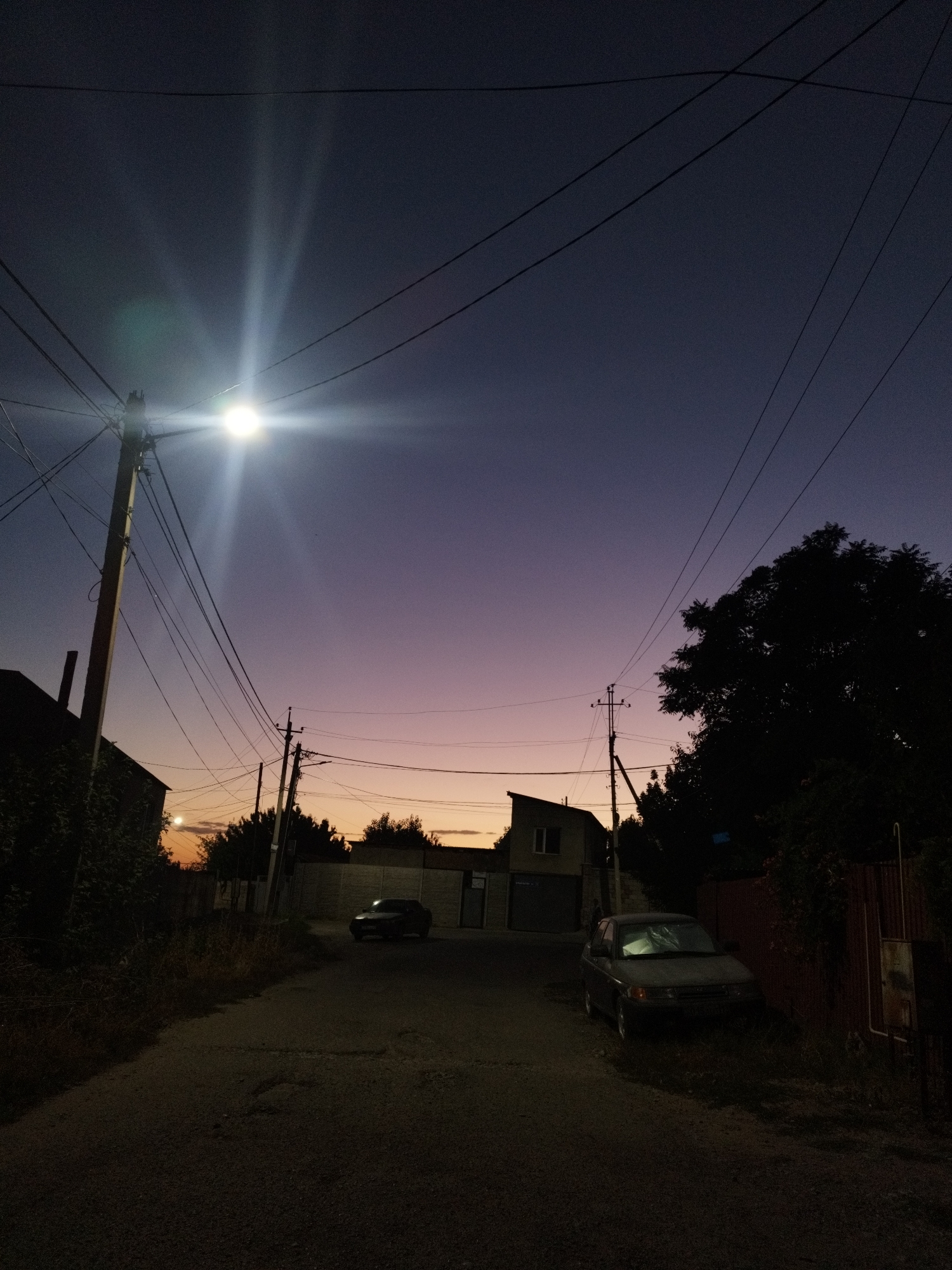 The image depicts a dusky street scene, likely in the city of Shymkent, Kazakhstan, as indicated by the context provided. The sky is a gradient of deep purple and soft pink hues, suggesting twilight. Power lines crisscross the frame, supported by wooden and metal poles, with a bright streetlamp illuminating the left side of the image, casting rays of light.
On the left side of the road, a dark-colored house with some greenery is visible. Further down the street, a building with a distinct angular roof is present, with garage doors. On the right side of the street, parked near a reddish-brown fence, is a dark-colored car. A large, silhouetted tree dominates the right side of the frame, its branches intricate against the fading light. The road itself appears to be asphalt, with some visible cracks and texture. The overall impression is that of a quiet, residential street at the end of the day.
