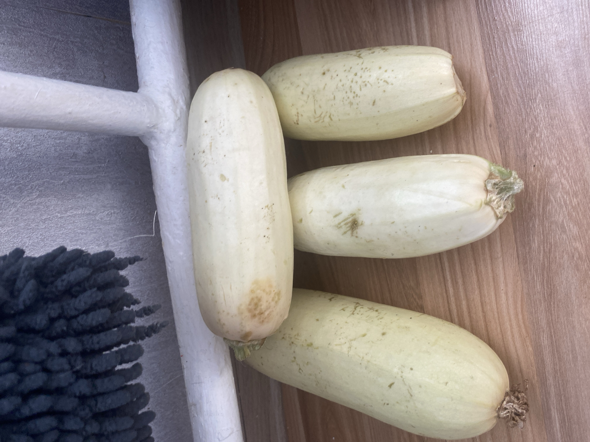 Four light green zucchinis are arranged on a wooden surface. Three are standing upright, side-by-side, with their stems at the bottom. The fourth zucchini is lying horizontally on top of the middle two. A white pipe runs across the top of the image, with a textured dark blue object hanging above it. The background appears to be a light gray textured wall.