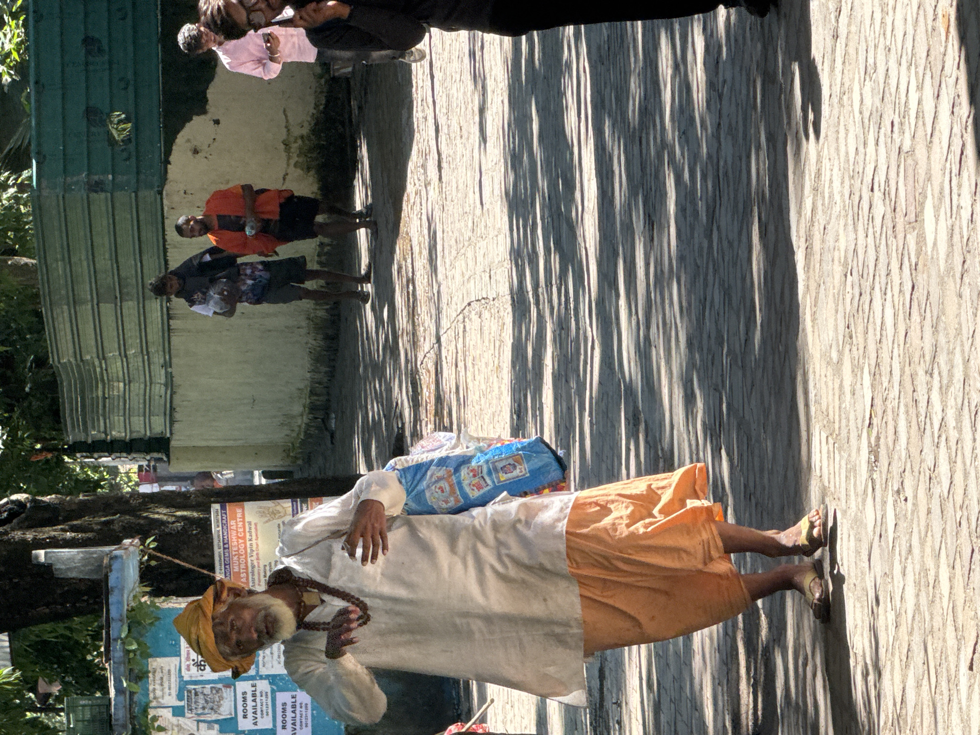 The image captures a street scene in Rishikesh, India, characterized by a paved pathway with brickwork and dappled sunlight casting long shadows. In the foreground, an elderly man with a white beard, wearing a yellow turban, a white tunic, and orange lower garments, walks towards the viewer. He has a mala (beaded necklace) around his neck and carries a blue and white patterned bag over his right shoulder. His left hand is raised, and his right hand is touching his chest. Behind him, two younger men are visible walking away from the camera. One wears a dark shirt and shorts, carrying a clear plastic bag, and the other, in an orange shirt and dark shorts, holds a water bottle. Further in the background, another person in a pink shirt is seen walking away, and to the far right, a person in dark clothing is partially visible. The background features a cream-colored wall, a green metal fence, and trees, suggesting an outdoor urban environment. Posters are visible on a board to the left of the main subject.