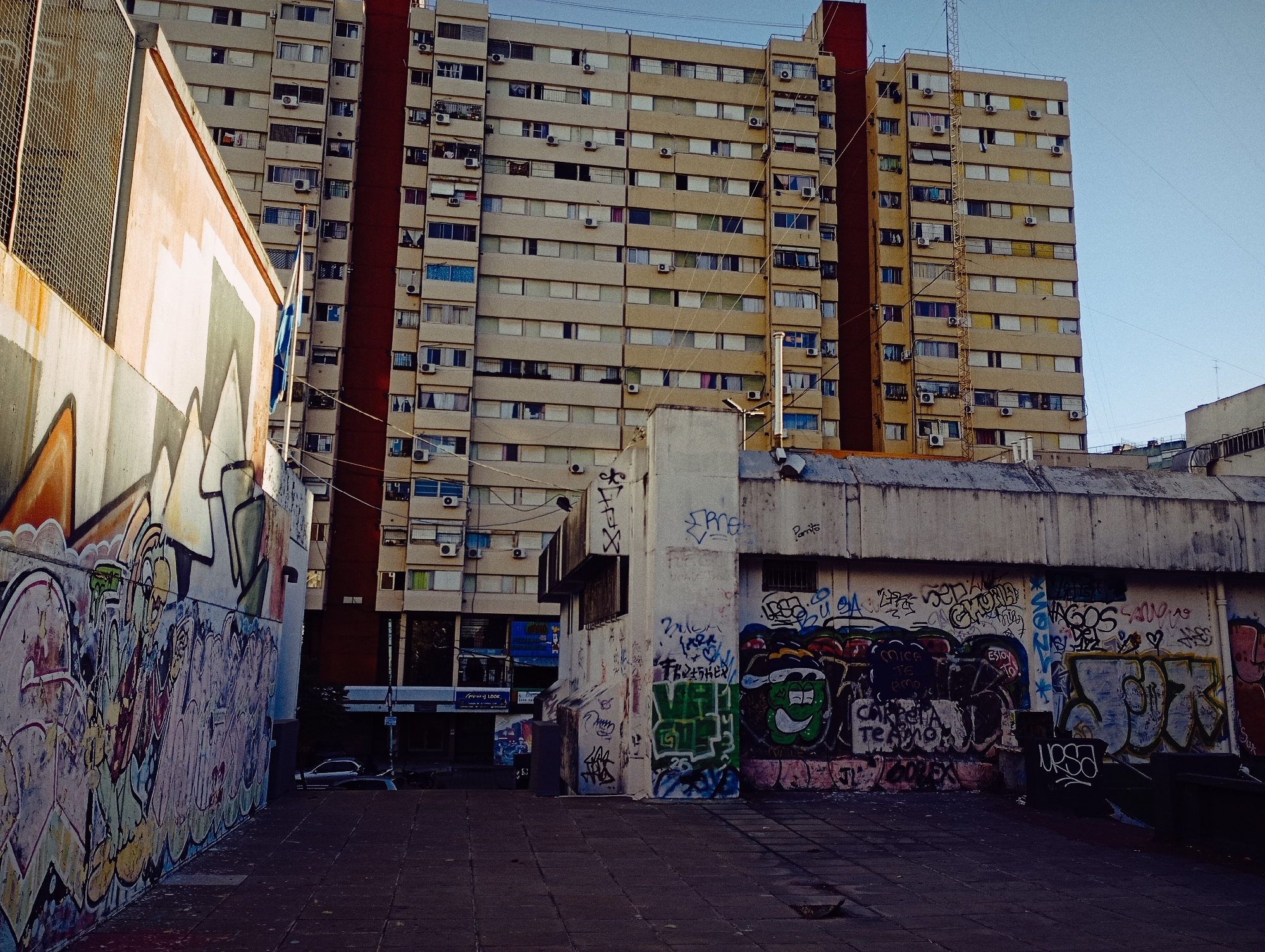 The image depicts an urban scene in Buenos Aires, Argentina, characterized by a large, multi-story apartment building in the background. The building is mostly beige with two vertical red stripes on its face and numerous windows, many with air conditioning units. In the foreground, there are walls covered in extensive graffiti. The walls flank a paved area, possibly a plaza or alleyway. A glimpse of a street with a few parked cars can be seen between the graffiti-covered walls. The sky is visible and appears to be a clear blue. There are no visible people in the image. The scene has an overall gritty and urban atmosphere.