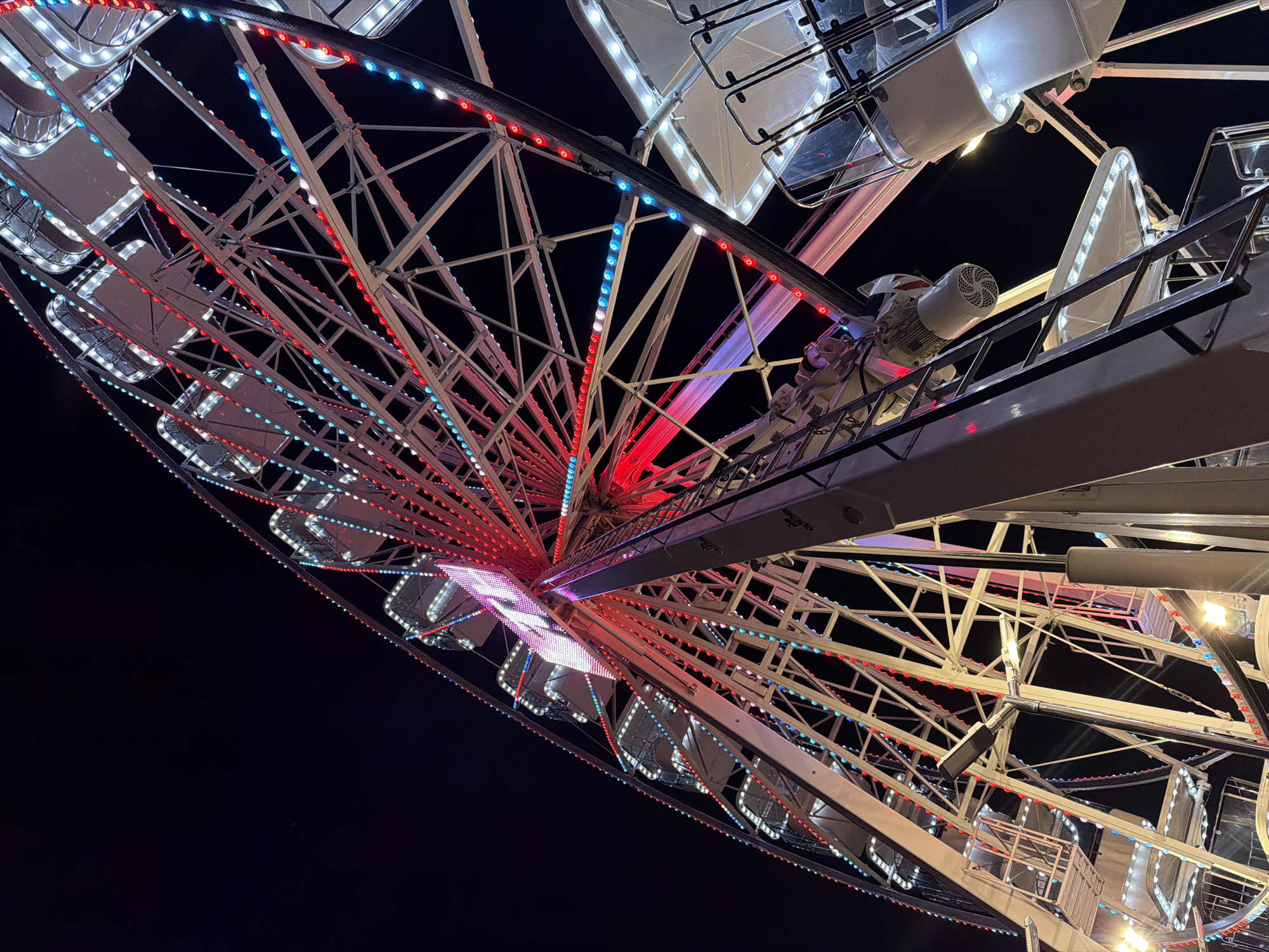 This is a low-angle, nighttime photograph of a Ferris wheel illuminated with red, white, and blue lights. The Ferris wheel is a large, circular structure made of metal scaffolding and is adorned with numerous small lights that outline its spokes and cabins. Several cabins, appearing white with black frames and clear protective panels, are visible, some containing passengers. A ladder extends vertically along the central support structure of the Ferris wheel. The background is dark, indicating it is nighttime. The scene appears to be an amusement park or fairground. The Ferris wheel is located in Gatteo, Italy.