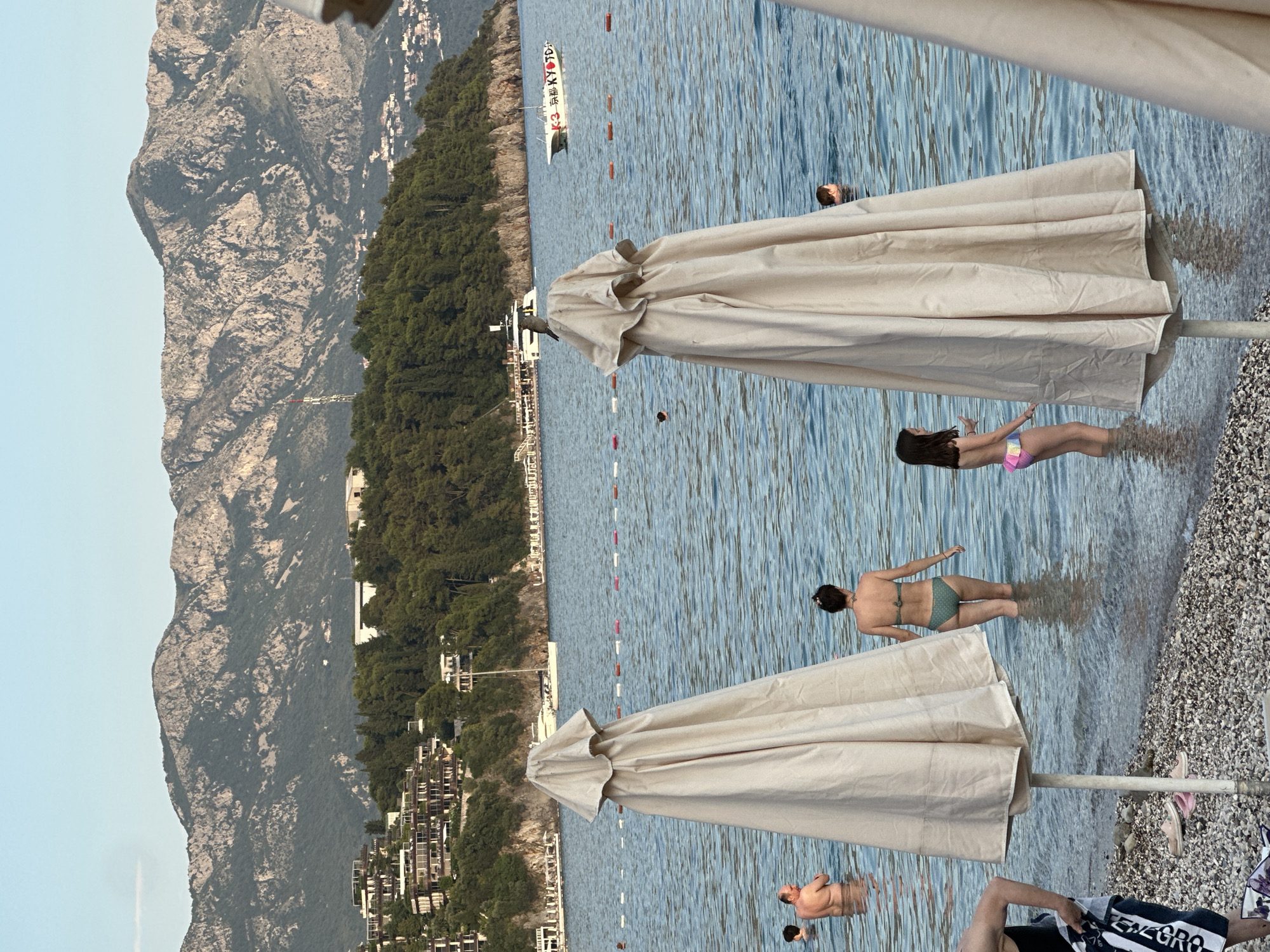 The image depicts a coastal scene in Budva, Montenegro, likely on a beach. In the foreground, two large, cream-colored beach umbrellas are prominently positioned. The one on the left is closer to the viewer, obscuring part of the water. The one on the right is slightly further away. The beach itself is covered in small grey and white pebbles.
In the water, several people are visible. To the left, a man is standing and looking out at the sea. Further left, another person is partially visible in the water. A woman wearing a green bikini stands in the shallow water, facing away from the camera. To her right, a young girl in a colorful bikini also stands in the water. Another person's head is visible further out in the sea.
The water is calm and blue, with a row of red and white buoys marking a boundary in the distance. Beyond the water, a lush green hillside slopes upwards, covered in trees. Dotted along the hillside are modern buildings, appearing to be resorts or hotels with white facades and multiple levels.
Dominating the background are large, rugged mountains with rocky grey peaks and green vegetation. The sky above is a clear, pale blue, suggesting it is late afternoon or early evening. A white boat with red lettering is visible in the water near the shore, and a larger white boat or yacht is anchored further in the distance.