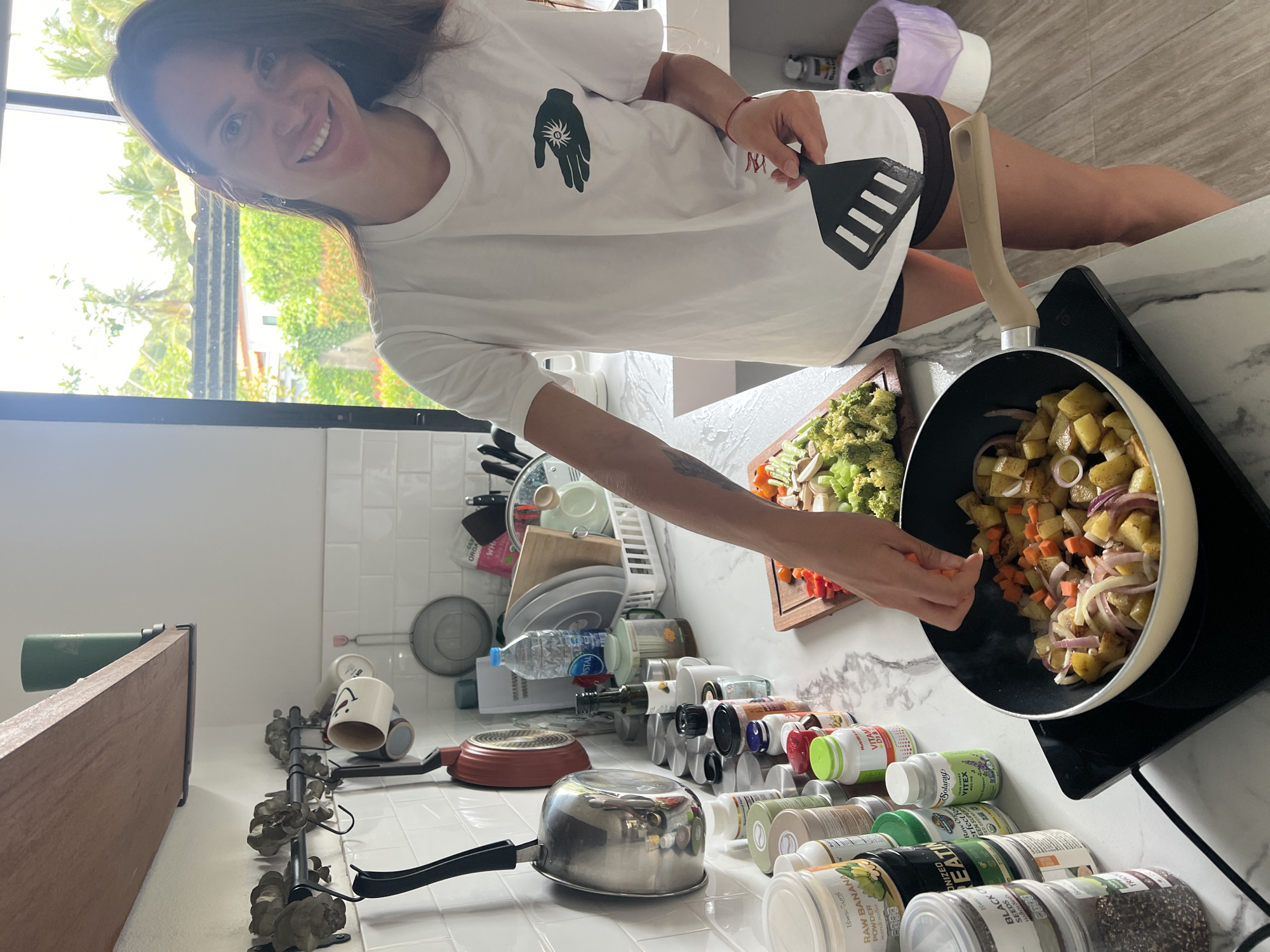 A woman is standing in a kitchen in Ban Bang Rak, Thailand, cooking. She is wearing a white t-shirt and black shorts, and is holding a spatula. In front of her, a pan on a stovetop contains potatoes, onions, and carrots. To the left of the stovetop are various bottles and containers, possibly supplements. To the right of the pan is a cutting board with chopped broccoli and red bell peppers. The kitchen has white tiled walls and a marble countertop. The background shows a window with lush green trees and vegetation visible, suggesting a tropical environment.