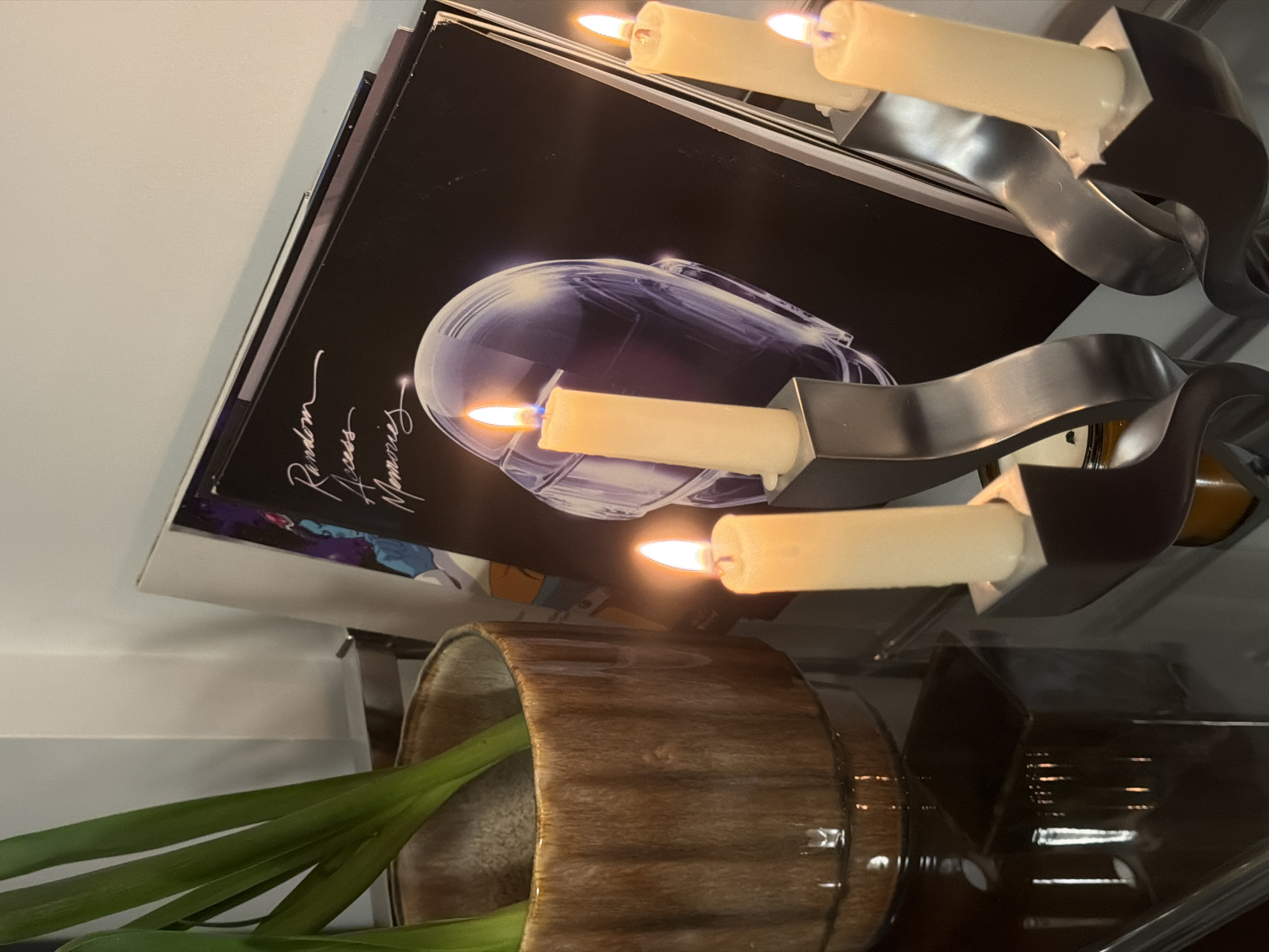 The image shows a close-up of a shelf with several items. On the shelf is a vinyl record album tilted towards the viewer, with the title "Random Access Memories" visible on the cover. Next to the album is a decorative candelabra with lit candles, and in front of the album is a brown pot containing green plants. The objects sit on a reflective surface. There is no visible context to indicate location in Montreal, Canada.