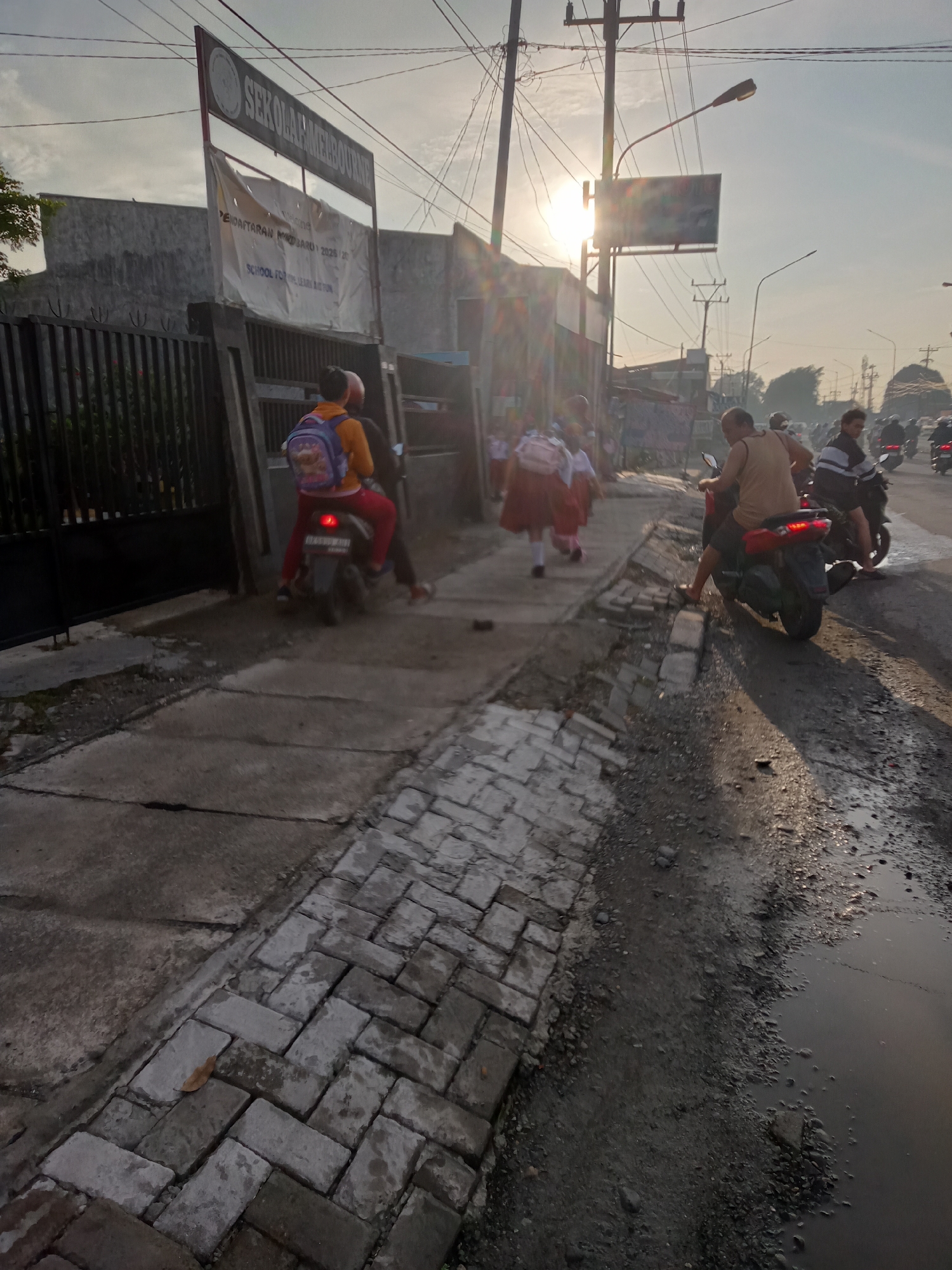 This image depicts a street scene in Puji Mulyo, Indonesia, likely in the morning given the low sun angle and hazy sky. A school sign, reading "SEKOLAH..." is visible on the left, with a banner below it that appears to be an announcement for registration. Several schoolchildren in uniforms are seen walking and riding on motorcycles. On the sidewalk, two children are on a motorcycle, one in a yellow shirt and red pants with a backpack, and another behind them. Further down the sidewalk, a group of schoolchildren in red and white uniforms are walking, suggesting they are heading to or from school. The street itself is a mix of asphalt and dirt, with puddles of water reflecting the sky. Several motorcycles are present on the road, with riders. The overall environment appears to be a typical urban or semi-urban street in Indonesia, with a mix of concrete structures, fences, and overhead electrical wires.
