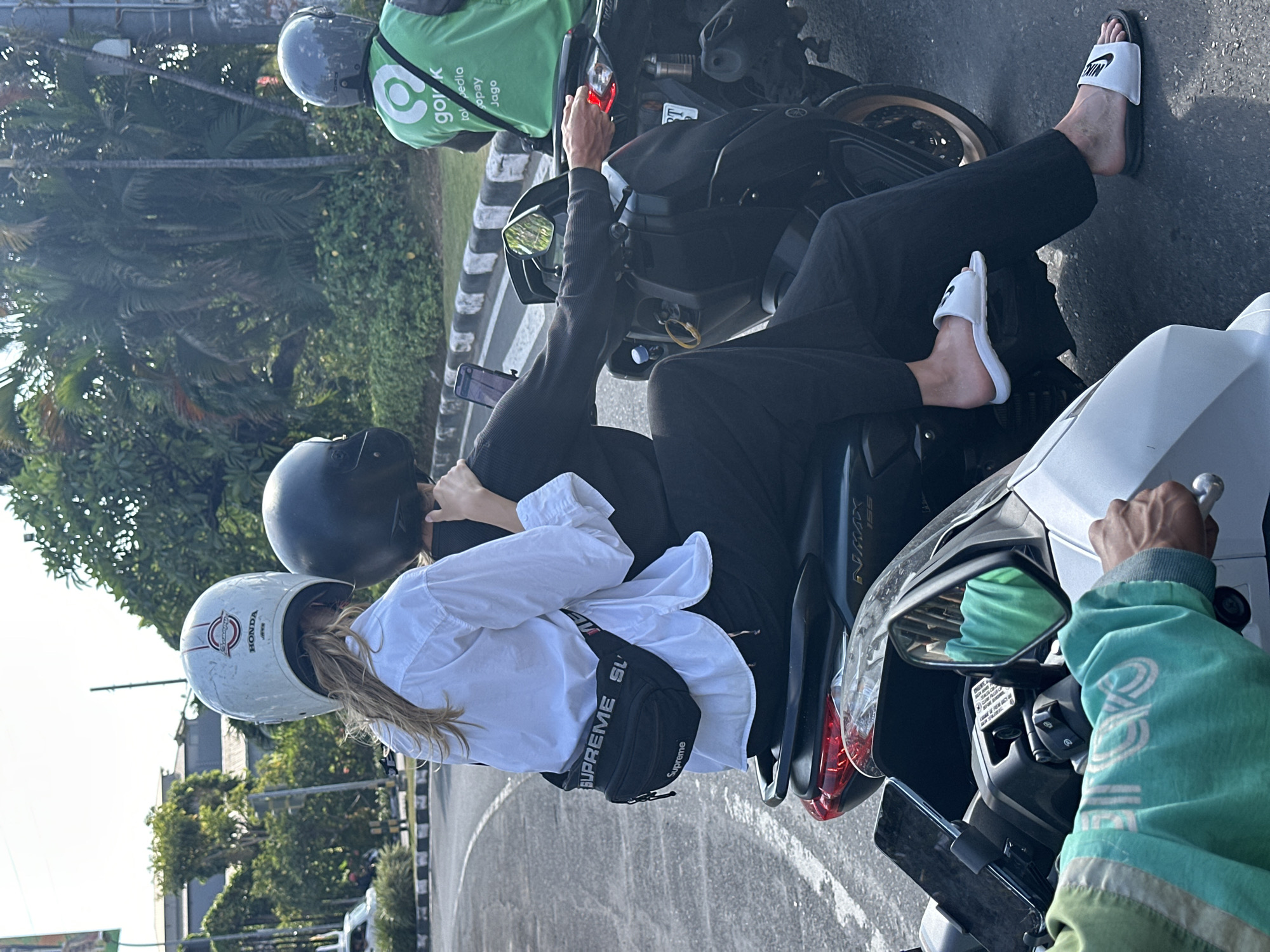 Two individuals are riding on a motorcycle in the city of Kuta, Indonesia. The person in front, wearing a white button-down shirt and black pants, is seated as the passenger. They have a white helmet with red and white stripes and a black "Supreme" fanny pack strapped across their waist. The person in the back, who is driving, is wearing a black helmet and black clothing. Both individuals are wearing sandals.
In the background, another motorcycle rider wearing a bright green vest with the "GoJek" logo is visible, also wearing a helmet. The motorcycles are on a paved road with a distinct white and black striped curb. Lush green tropical foliage and palm trees are present on the right side of the road, indicating a warm climate. The scene appears to be an intersection or a point where traffic is stopped, given the presence of multiple vehicles. The lighting suggests it is daytime.