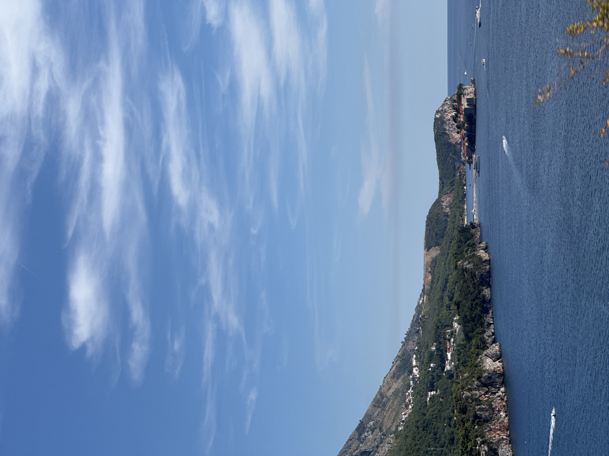 The image captures a bright, clear day with a vivid blue sky dotted with wispy white clouds. The scene overlooks a coastal landscape in Kamenovo, Montenegro, featuring a body of deep blue water. On the left side of the image, a steep, green-covered mountain slopes down towards the sea, with white buildings scattered along its side. Rocky cliffs jut out into the water in the foreground. Further out in the water, a small island is visible, densely populated with red-roofed buildings and fortified walls, connected to the mainland by a narrow causeway. Several boats, including speedboats and larger yachts, are present on the water, leaving white wakes behind them. The overall impression is one of a serene and scenic coastal vista.