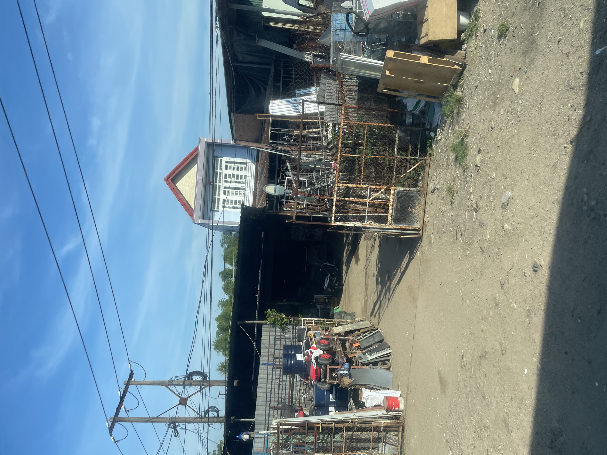 The image depicts a cluttered outdoor scene in what appears to be a workshop or salvage yard in Tay Nha Trang Ward, Vietnam. The foreground is a dirt and gravel lot, partially shaded by an unseen object. Several metal fences, some rusted, are present, one on the left and one on the right, with one of them open. Beyond the fences, there is a covered area with a dark roof, possibly a workshop. Inside this covered area, there are various objects including barrels, a red and white toy car, metal sheeting, and what looks like a motorcycle parked in the back.
Further in the background, a two-story building with a light blue facade and a red-tiled roof is visible, suggesting it is a residence or office. The sky is clear and blue with some wispy white clouds. Power lines are strung across the upper portion of the image, supported by wooden poles with metal crossbars. The overall impression is one of an active but unkempt industrial or recycling area, typical of some urban fringe environments. There are no people visible in the image.