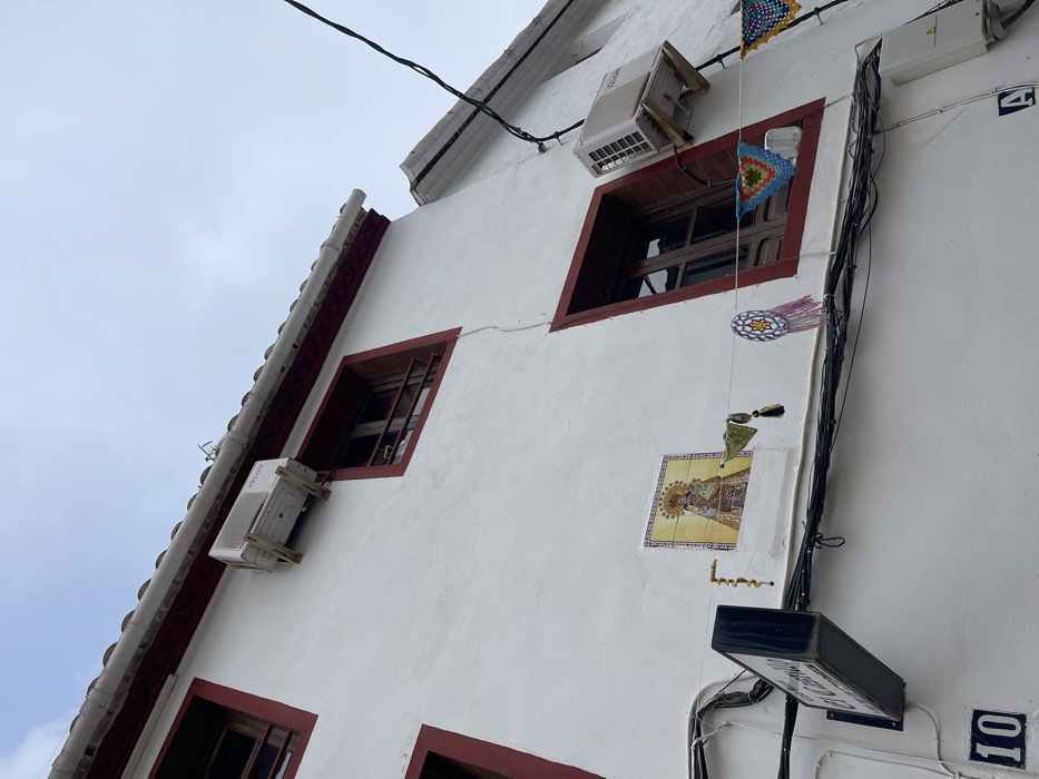 A low-angle exterior view depicts the white-stucco facade of a multi-story building in Buñol, Spain, likely near "El Castillo" as indicated by a prominent street sign. The building features dark red trim around its windows and along the roofline, which is topped with white, possibly terracotta, tiles. A light grey, overcast sky occupies the upper portion of the frame.
Three visible rectangular windows possess dark brown wooden frames; one in the upper left, one in the upper right, and a larger, open window in the mid-right. Two white Kosner brand outdoor air conditioning units are mounted on the facade with wooden brackets; one is positioned above the upper-right window, and another is to the right of the mid-right window. A vertical hairline crack extends down the white wall between the upper and lower sections.
Centered on the lower-mid facade is a rectangular ceramic tile mosaic depicting a crowned and haloed religious figure, likely the Virgin Mary, holding a child, set against a golden background with a yellow border. Below this mosaic, on the left, a dark-colored, rectangular sign bearing the white text "El Castillo" extends from the wall. The house number "10" is affixed below this sign, and the letter "A" is visible further right, below a dense bundle of black utility cables that traverse the facade horizontally. Various decorative elements hang from these cables and window frames: a string of light-colored beads to the left of the tile mosaic, a green crocheted triangular element with tassels, a circular multi-colored (purple, white, blue) crocheted ornament with tassels, and a blue, green, and red crocheted square draped across the mid-right open window. Another yellow and green crocheted item is partially visible on the far right.