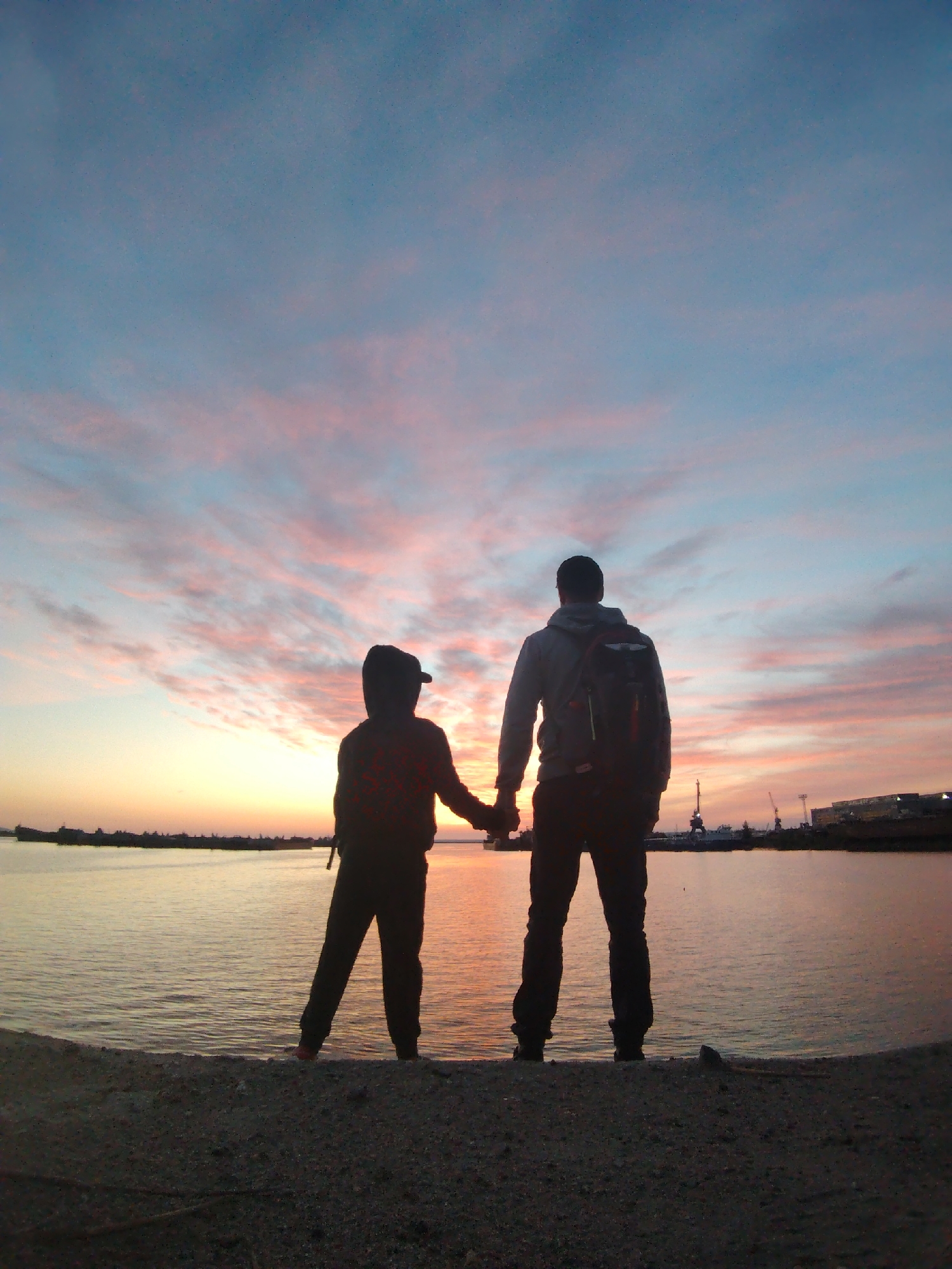 An outdoor photograph captures two individuals standing on a gravelly shore, facing away from the viewer, as they gaze out over a body of water during either sunrise or sunset. Both figures are rendered in silhouette against the vibrant sky. The person on the right appears to be an adult, wearing a hoodie and a backpack. To their left, a shorter figure, likely a child, wears a cap and a patterned shirt. They are holding hands, suggesting a shared moment.
The sky is a prominent feature, displaying a beautiful blend of warm colors like orange and yellow near the horizon, fading into soft pinks and light blues higher up, with delicate, wispy clouds scattered across. The water is calm, reflecting the fading light. In the distance, across the water, an industrial landscape is visible, characterized by the dark silhouettes of cranes, buildings, and what seem to be ships or boats, indicating a port or urban area. No visible text is present in the image. Given the location context, this serene yet industrially-tinged scene likely depicts the Kama River or Votkinsk Reservoir in Chaikovsky, Russia.