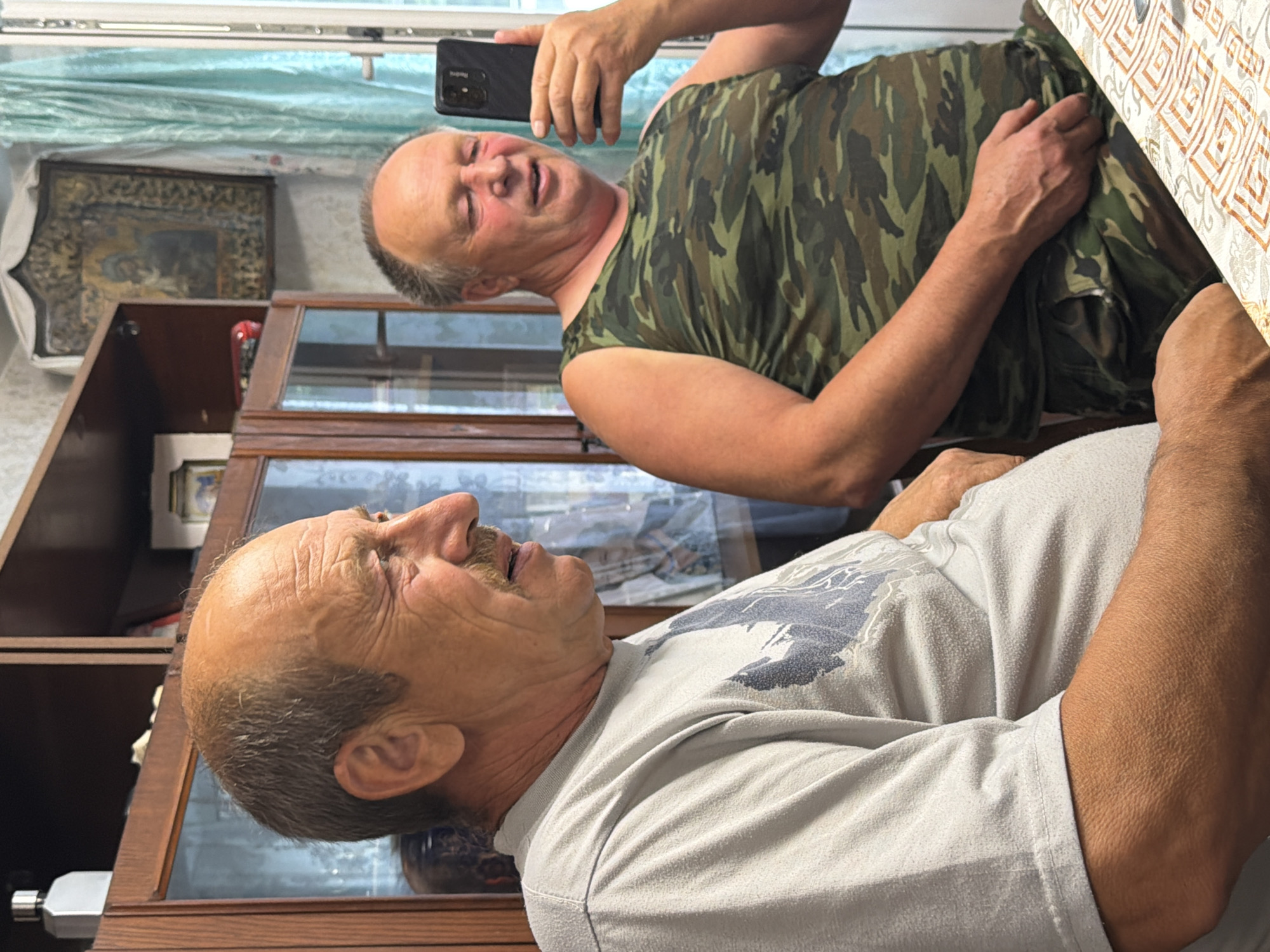 Two middle-aged men are seated at a table in what appears to be a residential interior. The man on the left is wearing a light gray t-shirt with a dark graphic print. He has a prominent mustache and is looking to his left with a slight smile. The man on the right is wearing a camouflage tank top and camouflage shorts. He is holding a smartphone in his right hand and appears to be taking a selfie or recording a video, looking at the phone's screen. The background shows wooden furniture, possibly a cabinet with glass doors, and a curtain with a teal hue. An icon or religious artwork is visible on the wall above and behind the man on the right. The table is covered with a patterned tablecloth. The lighting suggests an indoor setting, possibly during the daytime. The location is identified as Bronnitsa, Russia.