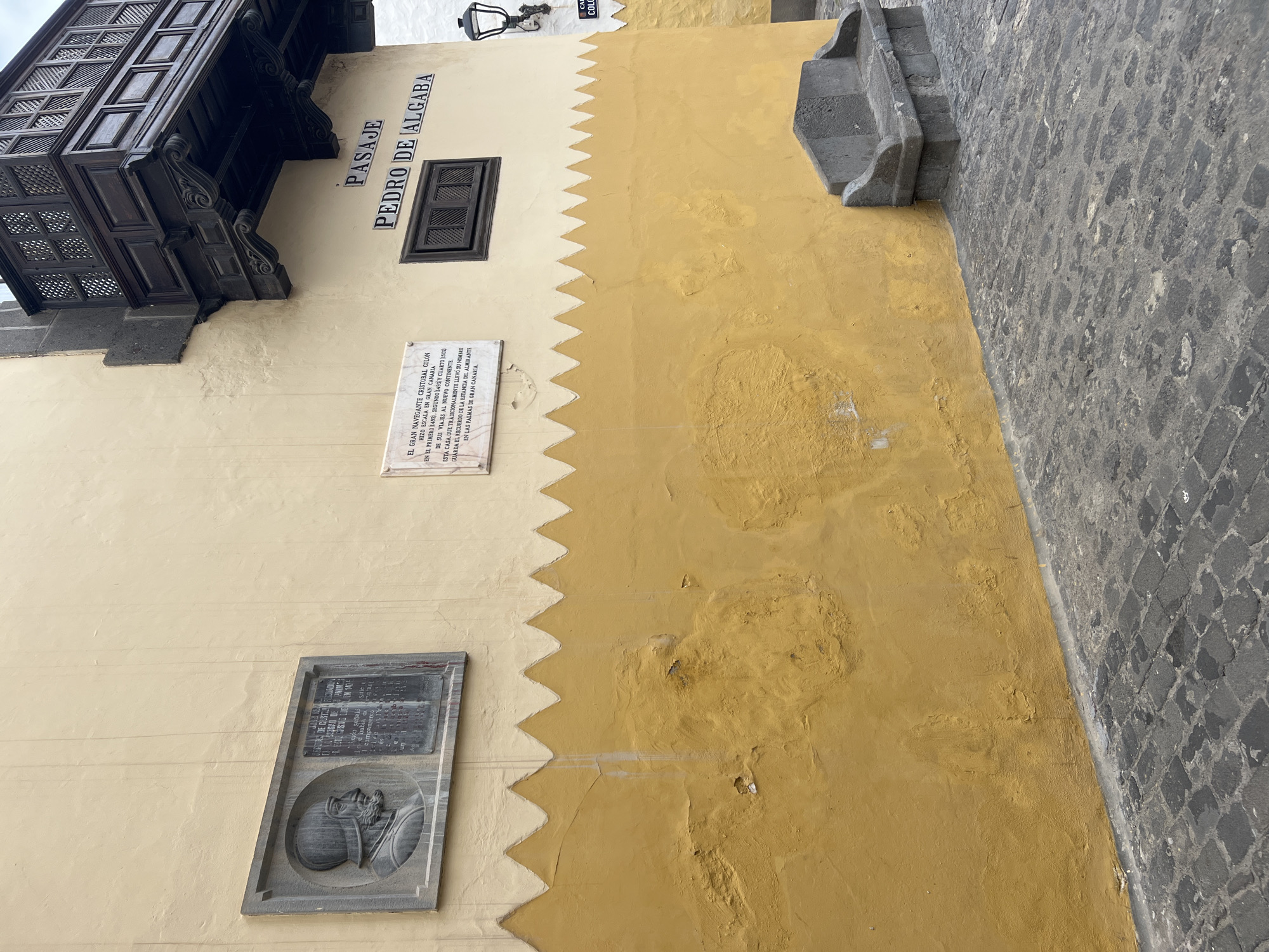 Here is a description of the image:
This is a ground-level shot of a building wall in Las Palmas De Gran Canaria, Spain. The wall has a decorative two-tone color scheme, with the top part being a lighter yellowish cream, and the lower portion being a darker mustard yellow. The demarcation line between these colors is zig-zagged.
The image features several plaques and architectural details attached to the wall:
1. A small, dark wooden window with a decorative exterior frame.
2. A black wooden balcony with intricate patterns of holes.
3. A white plaque with black lettering, containing historical information about Christopher Columbus' voyages to Gran Canaria.
4. A grey plaque featuring a relief of a person, possibly a historical figure or symbolic image.
5. A stone fixture extends from the wall, possibly a small balcony or architectural detail.
The words "PASAJE PEDRO DE ALGABA" are painted on the wall in black lettering, referring to the name of a local alleyway.
To the right of the wall, is a portion of the street, paved with grey cobblestones. The wall is clearly aged with some weathering and surface imperfections.
