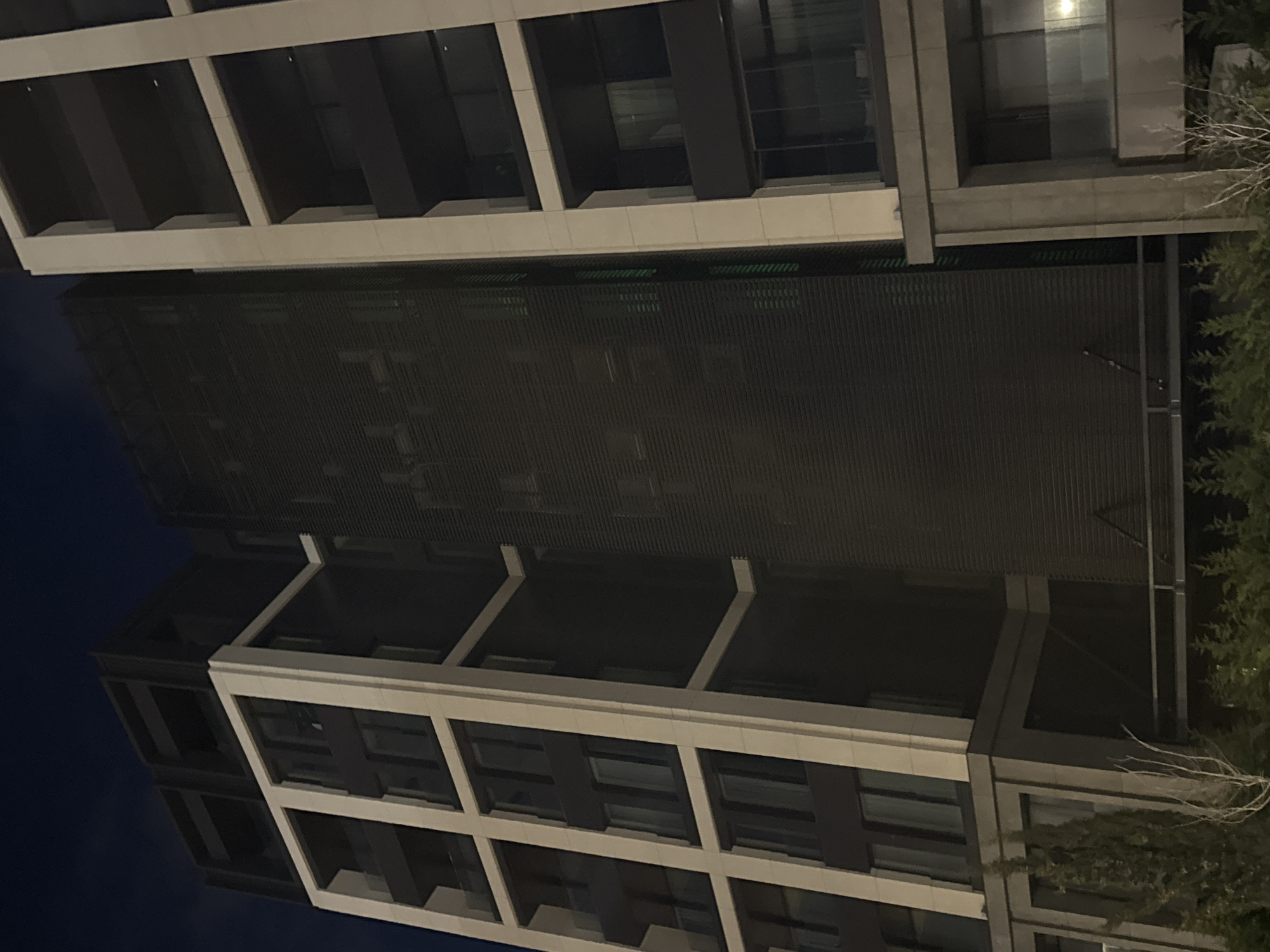 The image depicts an upward view of a modern building at night. The building features a repetitive pattern of windows and support structures, alternating between light and dark tones. A screen or awning-like structure projects horizontally from the building's midsection. The sky is dark blue. The building's construction suggests a contemporary architectural style. The location is unknown; there is no information in the image to identify Burgas, Bulgaria. There are no people, subjects, actions, or interactions visible. The environment is urban.