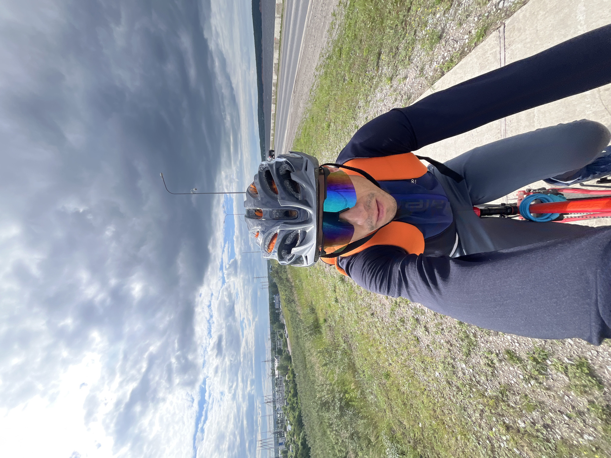 A man wearing a grey and orange helmet, blue and orange sunglasses, a dark blue long-sleeved shirt, and grey pants is riding a red bicycle on a paved path. He is also wearing an orange backpack. The man is looking at the camera. He is in the foreground of the image. In the background, there is a road with cars, a body of water, trees, and a cloudy sky. This scene is likely located in the city of Chaikovsky, Russia, as evidenced by the industrial structures and river visible in the distance, which are characteristic of the area around the Kama River.