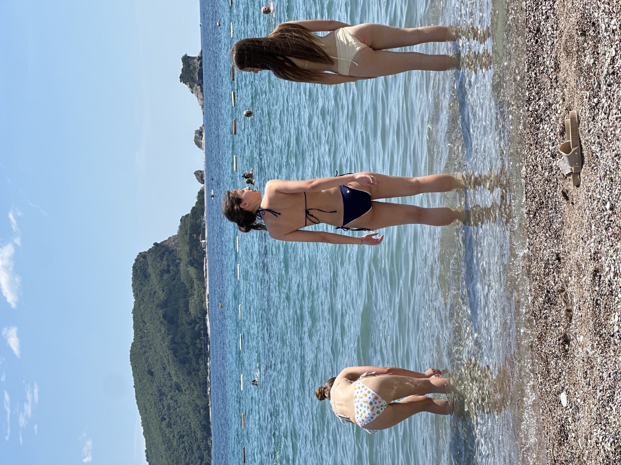 The image shows a beach scene in Budva, Montenegro. Three women wearing bikinis are wading in shallow water along the shoreline. The woman closest to the viewer is bent over, facing into the water. The woman in the middle is standing upright with her back to the camera. The third woman is further in the water facing into the water with her back to the camera. The water is clear and calm, reflecting the sunlight. The beach is pebbly. A pair of sandals is on the beach on the right side. In the background, there's a verdant hill and a blue sky with few clouds. Buoys are visible in the water.