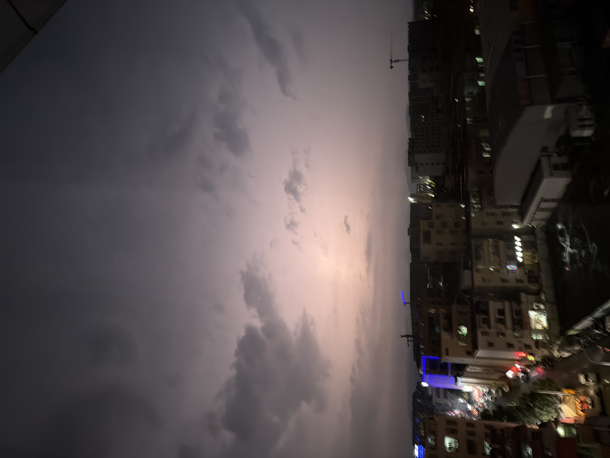 The image captures a city skyline at dusk or night, likely in Guangzhou City, China, as indicated by the context provided. The sky is predominantly filled with dark, ominous clouds, suggesting an approaching storm or recent rain. A faint glow emanates from the horizon, possibly due to the setting sun, city lights, or lightning within the clouds. The city below is a dense collection of buildings, ranging from multi-story residential blocks to commercial structures. Many buildings are illuminated with artificial lights, creating a vibrant, if somewhat blurry, urban landscape. A few cranes are visible, suggesting ongoing construction. The street level appears to be active with vehicles and illuminated storefronts, typical of a bustling city environment. The overall impression is one of a dynamic urban area under a dramatic sky.