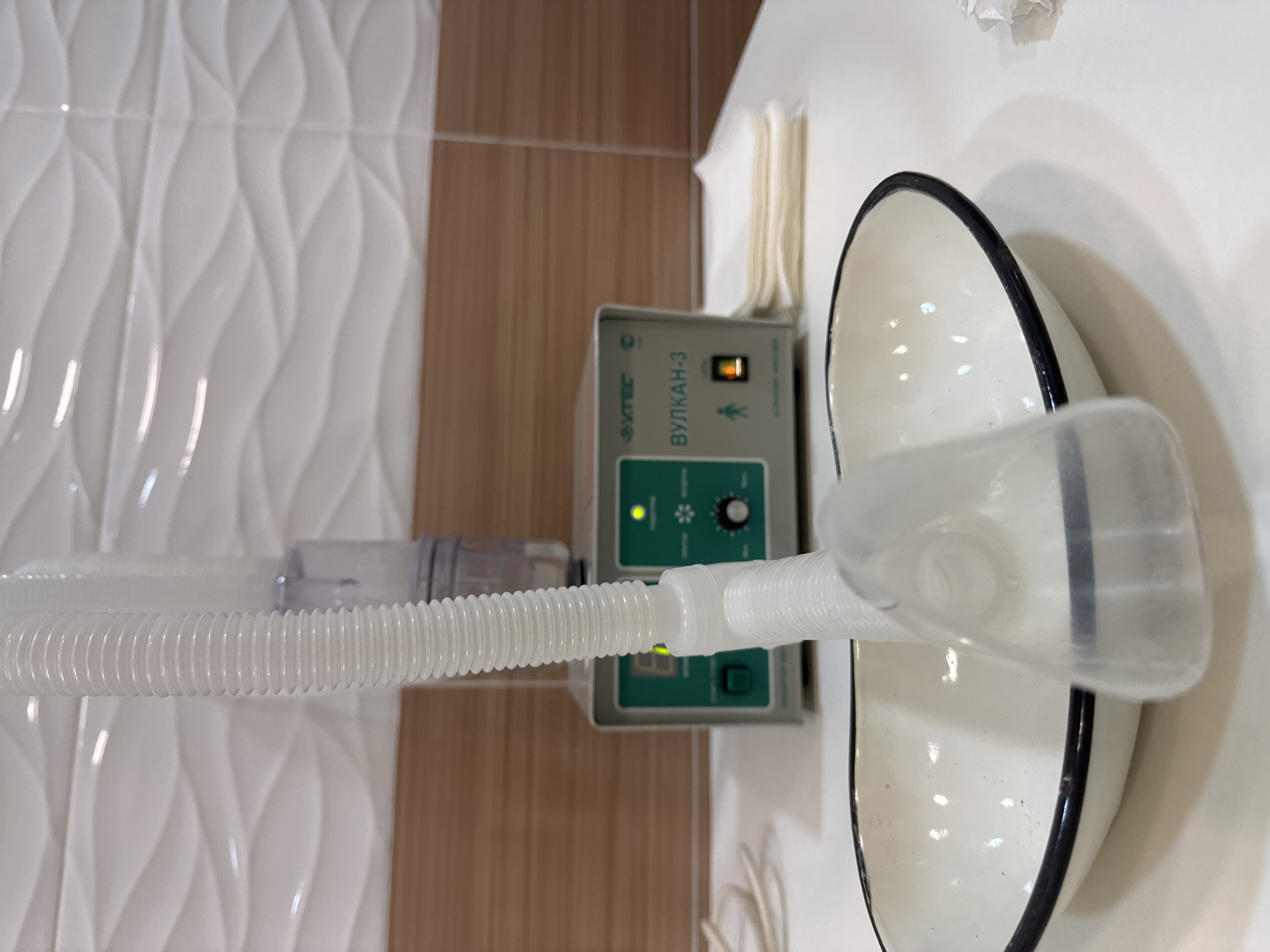 The image shows medical equipment, specifically an inhaler or nebulizer apparatus connected to a machine labeled "VULKAN-3." The machine is green and gray, with a control dial and indicator lights. A corrugated white tube connects the machine to a clear plastic mask, which rests inside a white bowl with a black rim. The background features white decorative tiles and wood-look tiles, suggesting the scene is located in a medical or treatment facility. There are also napkins or paper towels in the background. No people are visible. The text on the equipment indicates it is likely located in a Russian-speaking country, but there is no specific indication that it is located in the city of Chaikovsky.