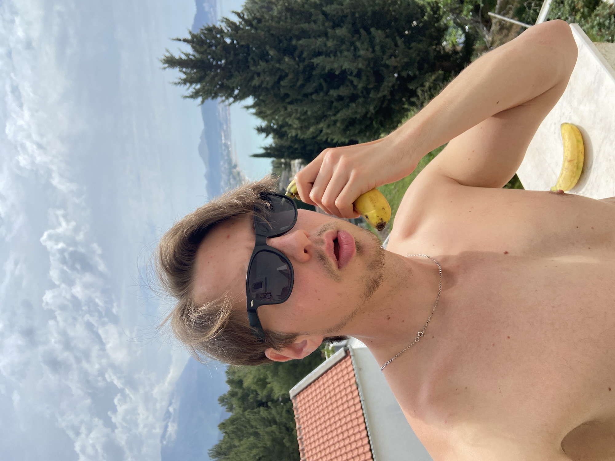 A shirtless fair-skinned male wearing sunglasses is holding a banana near his face. He has short brown hair and a stubbled beard. A silver chain necklace is visible around his neck. Another banana is lying on a white surface to his right. The background includes a view of the sky with clouds, trees, a body of water, mountains, and a building with a red tiled roof. This image appears to be taken outdoors in Šušanj, Montenegro, as the location matches the landscape features.
