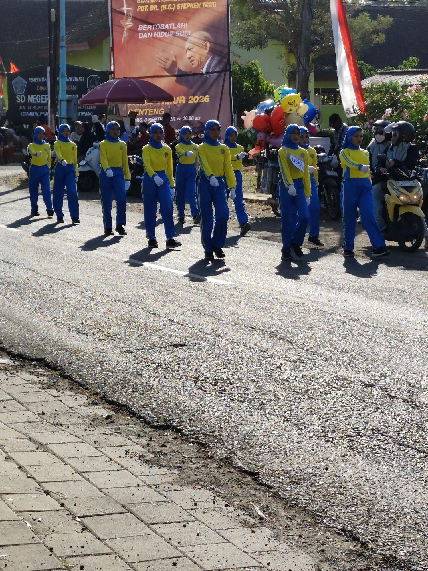 In the city of Sempu, Indonesia, a marching band composed of young students in bright yellow tops and blue pants proceeds down a street. The students are wearing blue headscarves. The students are walking in formation, with some holding their arms up. In the background, a large banner with Indonesian text and a picture of a man is displayed. A red and white Indonesian flag is also visible. Several motorcycles are parked on the side of the road, with some riders wearing helmets. Balloons are also visible, possibly part of the parade. The street is paved with asphalt, and there is a sidewalk made of concrete tiles on the left side of the image. The scene appears to be a public event or celebration.