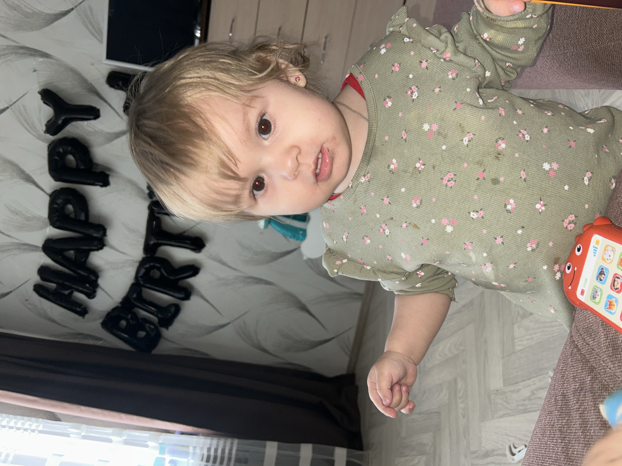 A young, fair-skinned child with blonde hair and brown eyes is the primary subject of the image. The child is wearing a long-sleeved, patterned top. There are smudges or stains visible on the child's face and clothes. An object appears to be in the child's hand, and a red toy phone is lying on a brown textured surface. In the background, black balloons spelling "Happy Birt..." are affixed to a wall with a wavy design. A dark-colored curtain or drape is visible at the bottom of the frame. The scene appears to be indoors, likely a child's birthday celebration based on the balloons. The image was captured in the city of Chaikovsky, Russia.