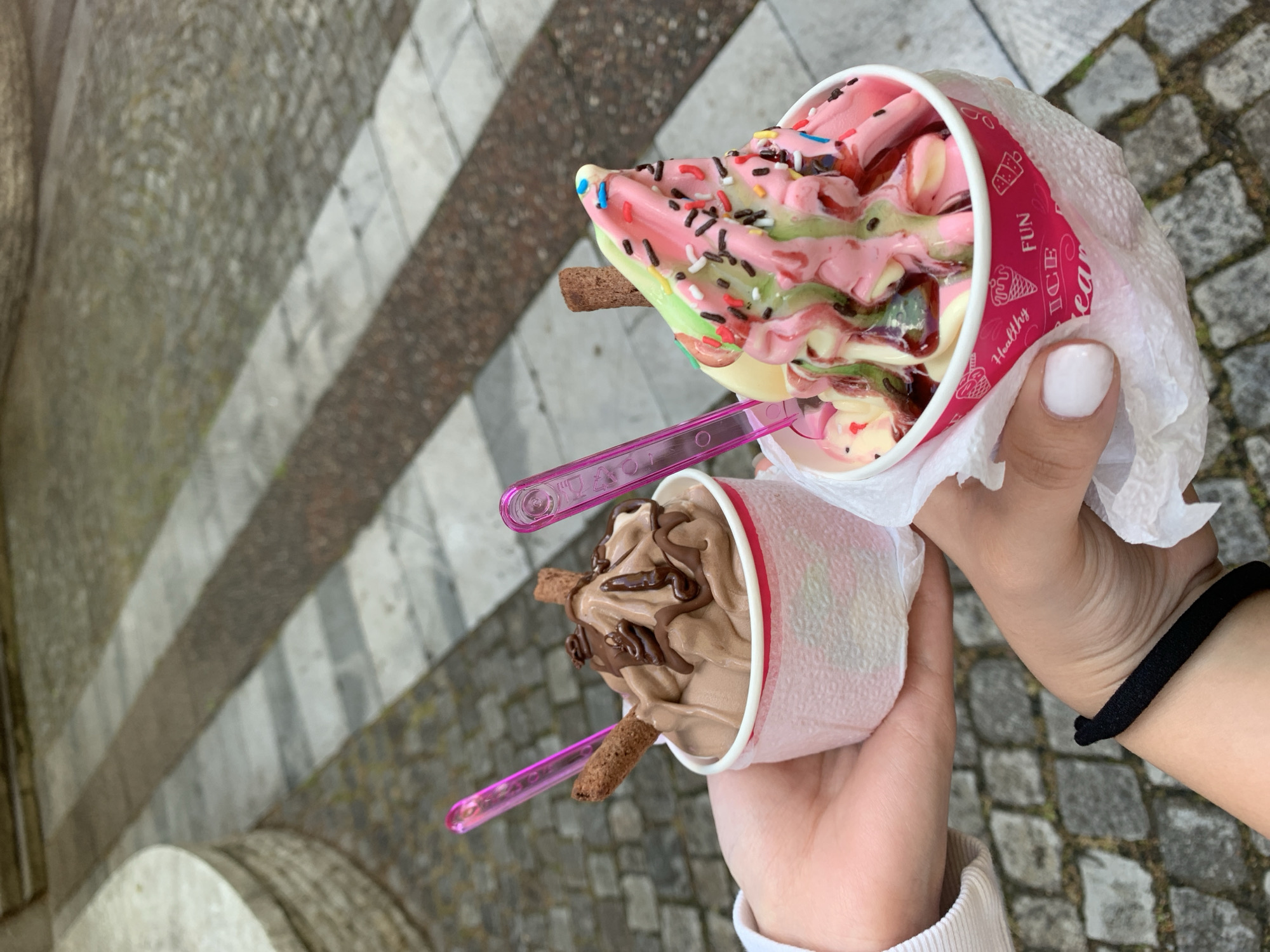 The image captures two hands holding cups of ice cream, viewed from an overhead, slightly tilted perspective, against a cobblestone background in what appears to be Kyustendil, Bulgaria.
The upper hand, featuring a thumb with white nail polish, holds a cup of multi-colored ice cream. This dessert showcases swirls of pink, light green, and a creamy yellow/vanilla, liberally topped with a mix of chocolate and colorful sprinkles. A chocolate wafer stick and a pink plastic spoon are embedded in the ice cream. The cup is partially wrapped in white tissue, beneath which a pink design is visible, featuring text such as "Healthy," "FUN," and "ICE CREAM," alongside small icons of ice cream cones and a cake.
The lower hand, with a black hair tie on its wrist, holds a cup filled with chocolate ice cream. This serving is artfully drizzled with chocolate syrup and garnished with two chocolate wafer sticks, also accompanied by a pink plastic spoon.
The background consists of a cobblestone pavement, suggesting an outdoor pedestrian area. Various striped patterns of grey and brown stone are visible, along with a curved stone structure in the bottom left, indicative of urban architecture. The lighting suggests it is daytime. The scene conveys a casual moment of enjoying desserts outdoors.
