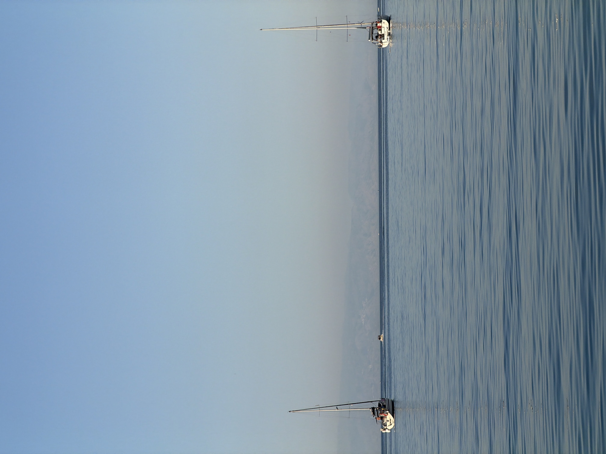 Two sailboats are sailing on a calm body of water under a clear sky. The location appears to be near Chianchitta, Italy, based on the coastal geography visible in the background. The sailboats are spaced apart with one in the foreground and the other in the distance. The water has subtle rippling patterns.