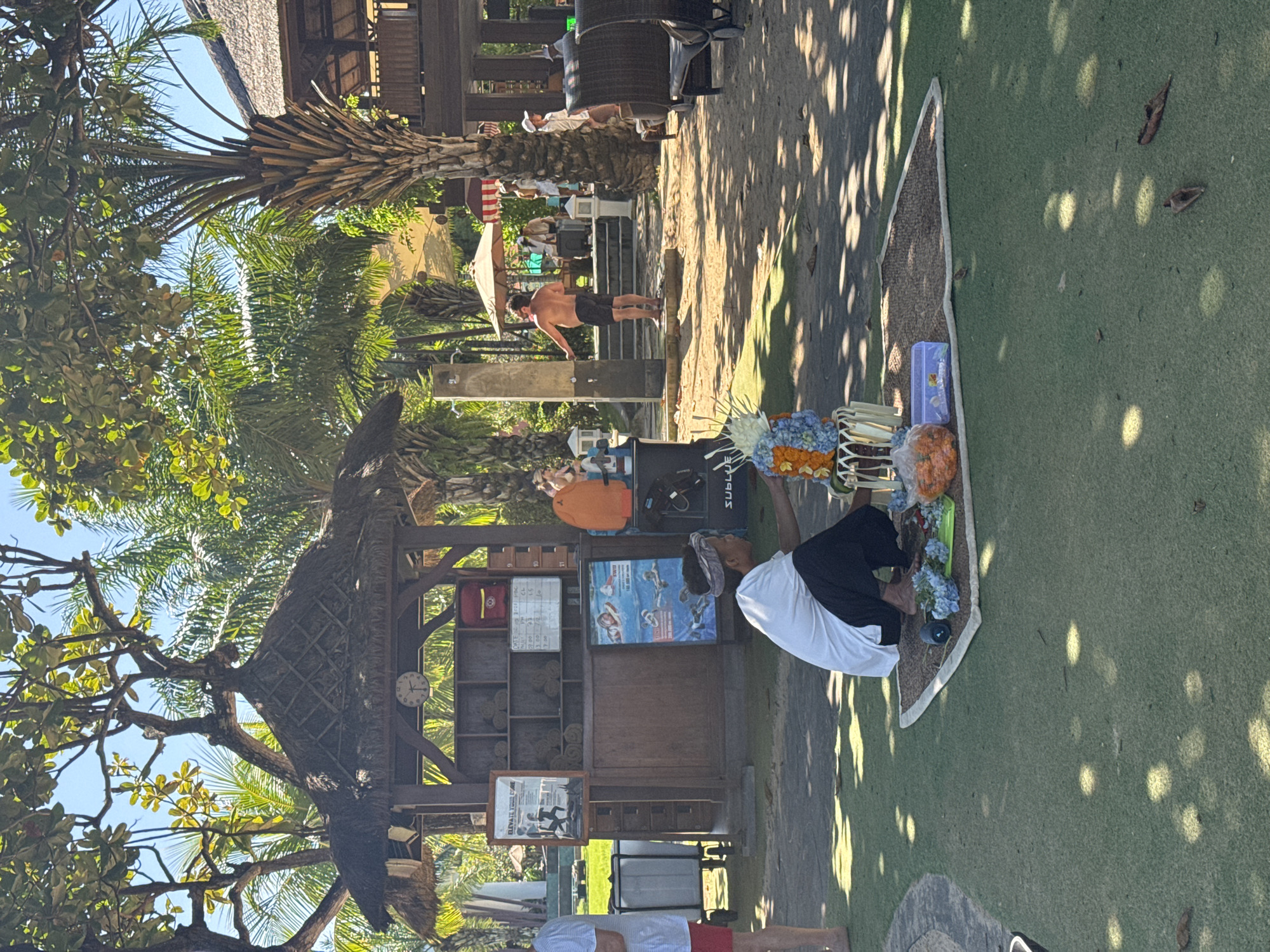 The scene is in Kuta Selatan, Indonesia, at an outdoor establishment. A man wearing a white shirt and dark pants is squatting near a display of flowers and items on a small rug or mat placed on green artificial turf. He appears to be arranging or tending to the floral display. Nearby is a wooden structure with shelves holding towels and signage. To the left, lush trees and plants create a canopy. In the background, people are milling about in a partially shaded area with tables and chairs. One man is using an outdoor shower. The ground is covered with shadows and sunlight patterns.