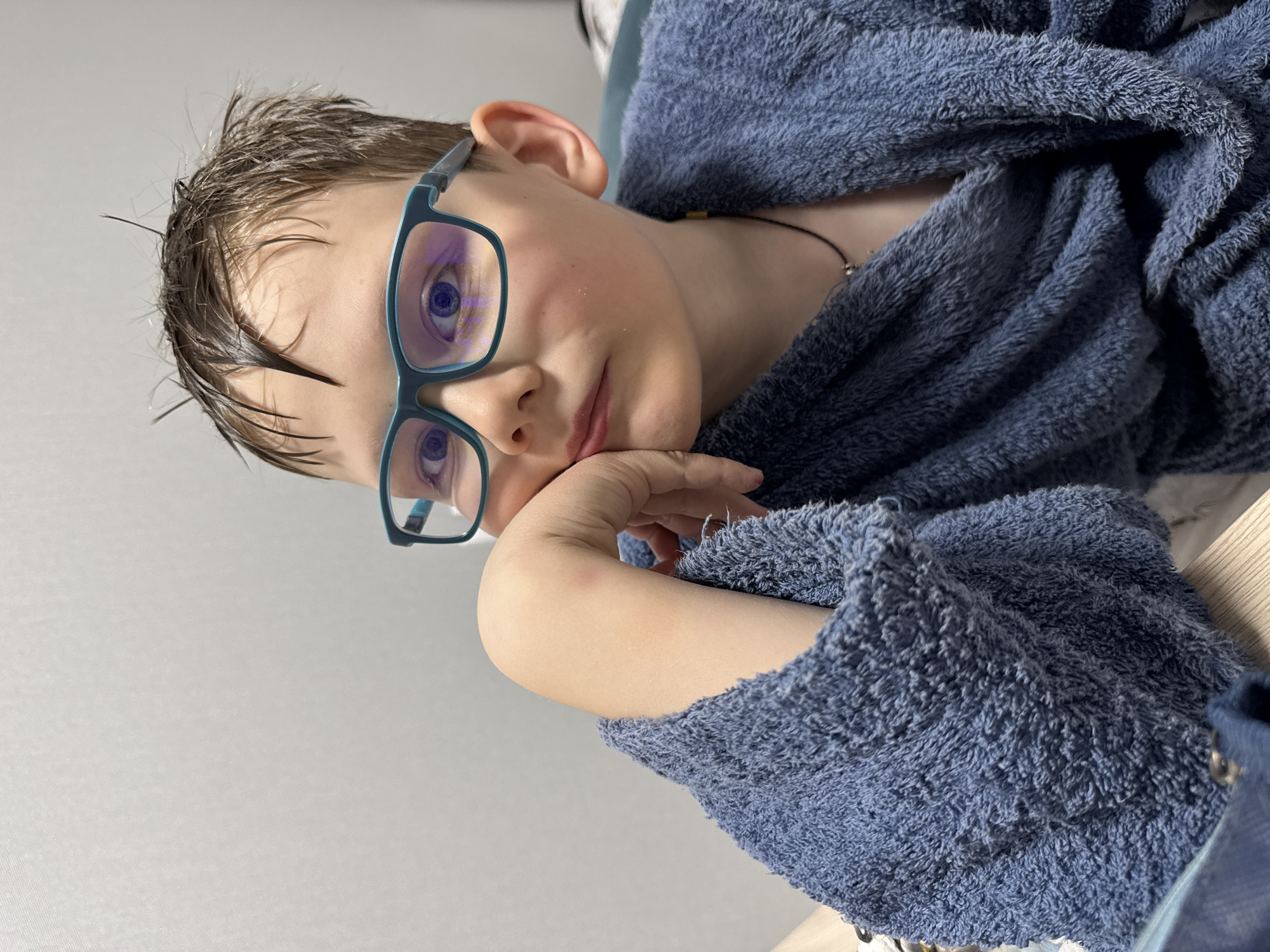 A young boy with wet hair and blue-framed glasses is resting his head on his hand. He is wrapped in a blue robe. The background is a plain, light-colored wall. The image seems to be taken indoors. The exact location in Gornyi (Dvurechenskoe S/P), Russia is unknown based solely on the provided image.