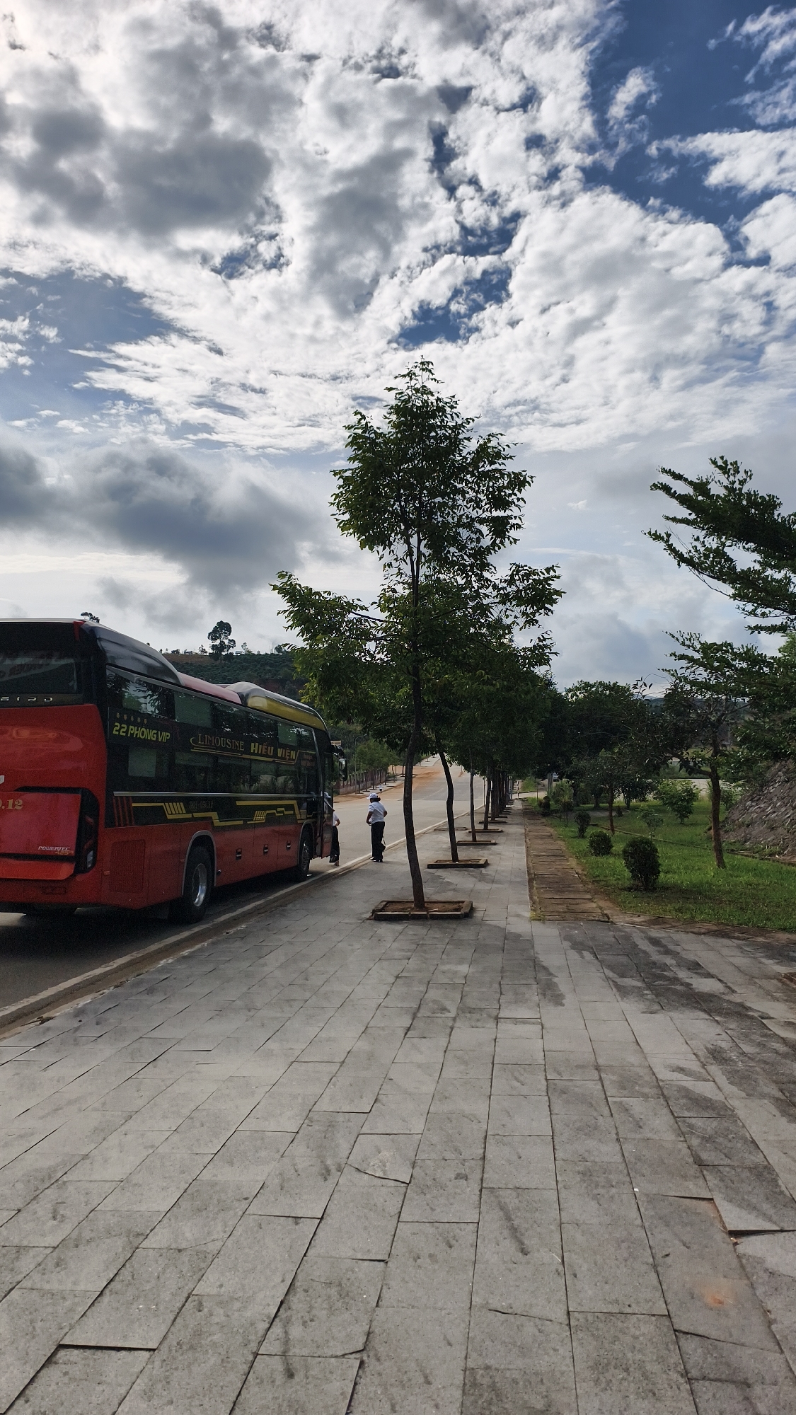A red bus with yellow and black accents is parked on the side of a paved road. A person in a white shirt and dark pants stands near the bus. The road leads to a sidewalk lined with young trees, which then transitions into a grassy area and a hillside covered in vegetation. The sky is cloudy.