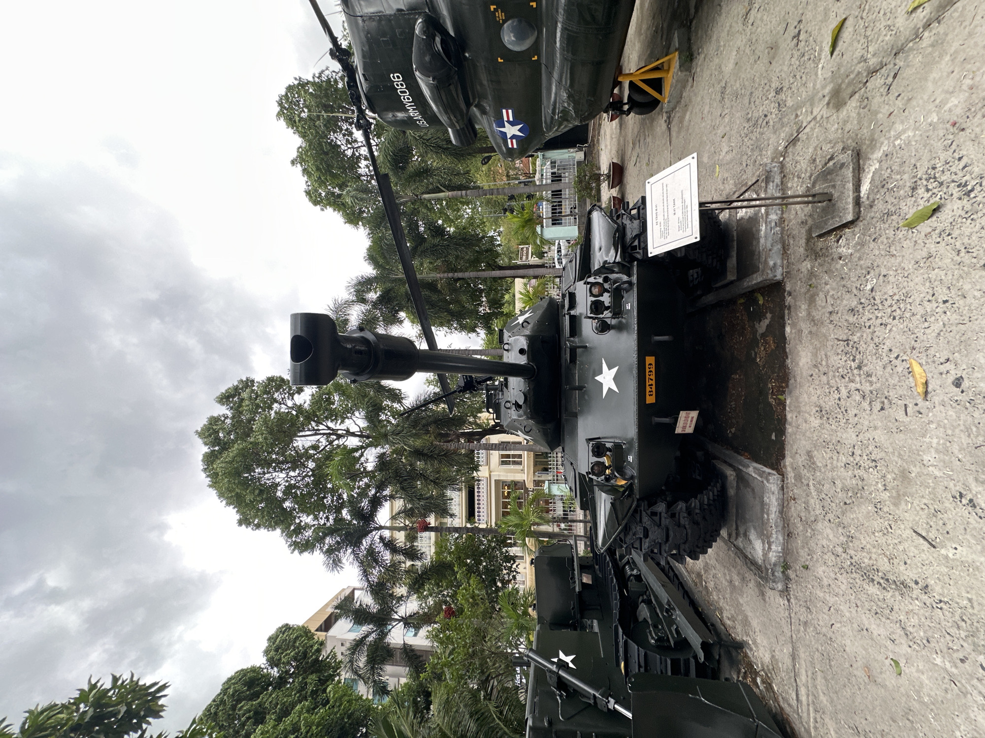 The image depicts an M41 Walker Bulldog light tank on display at a museum or memorial site. The tank is painted in military green with white stars as markings. In the background, there's a helicopter also in military green. The location appears to be outdoors, possibly a park or courtyard, in the city of Thủ Đức, Vietnam. Palm trees and other foliage are visible. Overhead, the sky is overcast. In the background, some buildings are also visible. A sign provides information about the tank. The ground is concrete.