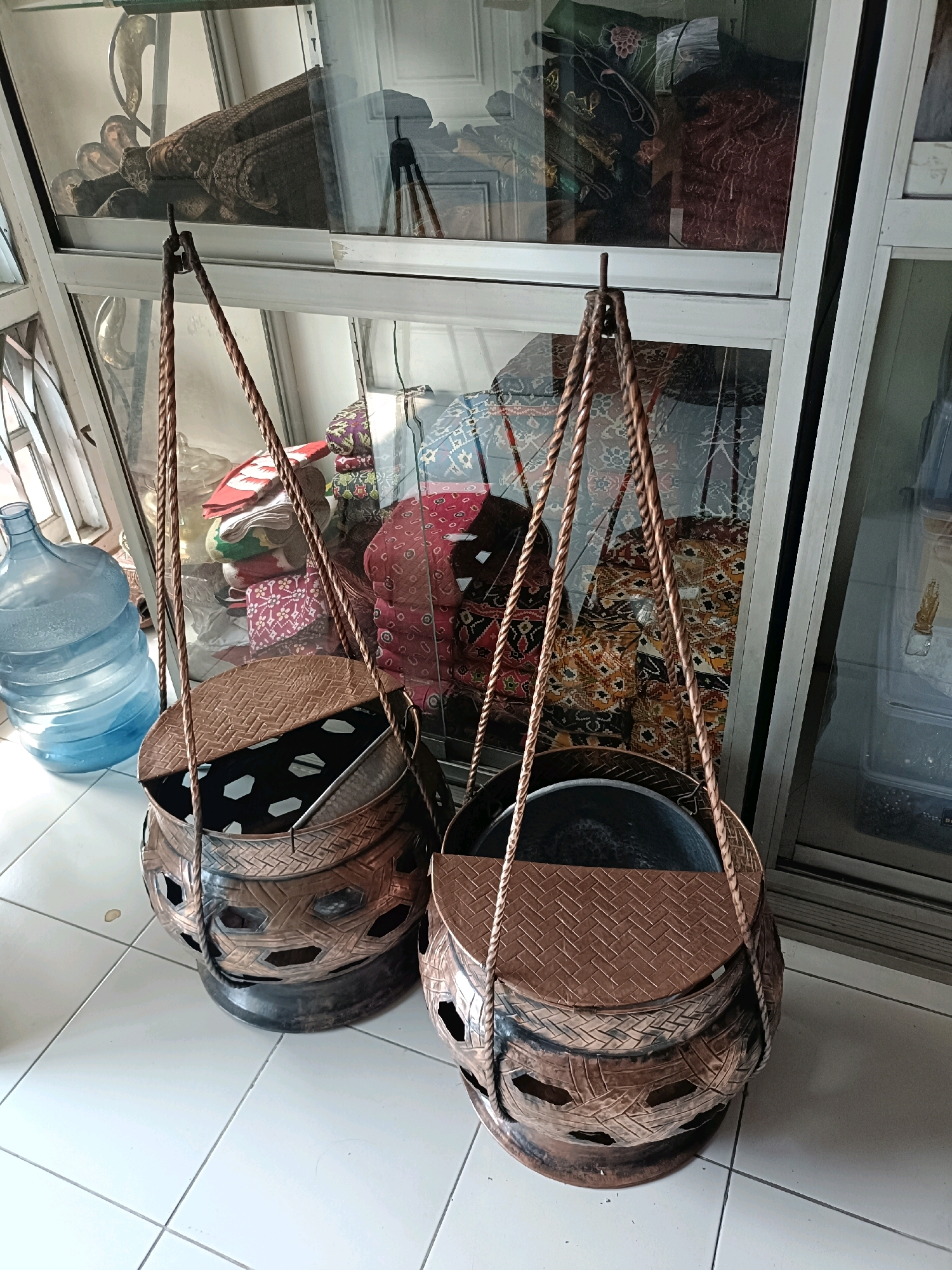Two ornate, copper-colored woven baskets with rope handles hang in front of a display case filled with colorful fabrics. The baskets are cylindrical with intricate cut-out patterns along their sides and woven lids. A large blue water cooler is visible to the left of the baskets. The display case behind them reflects a variety of fabrics, some with batik patterns. The scene appears to be set on a tiled floor. The image was taken in Semarang, Indonesia, suggesting a location such as a shop selling traditional Indonesian crafts or home goods.