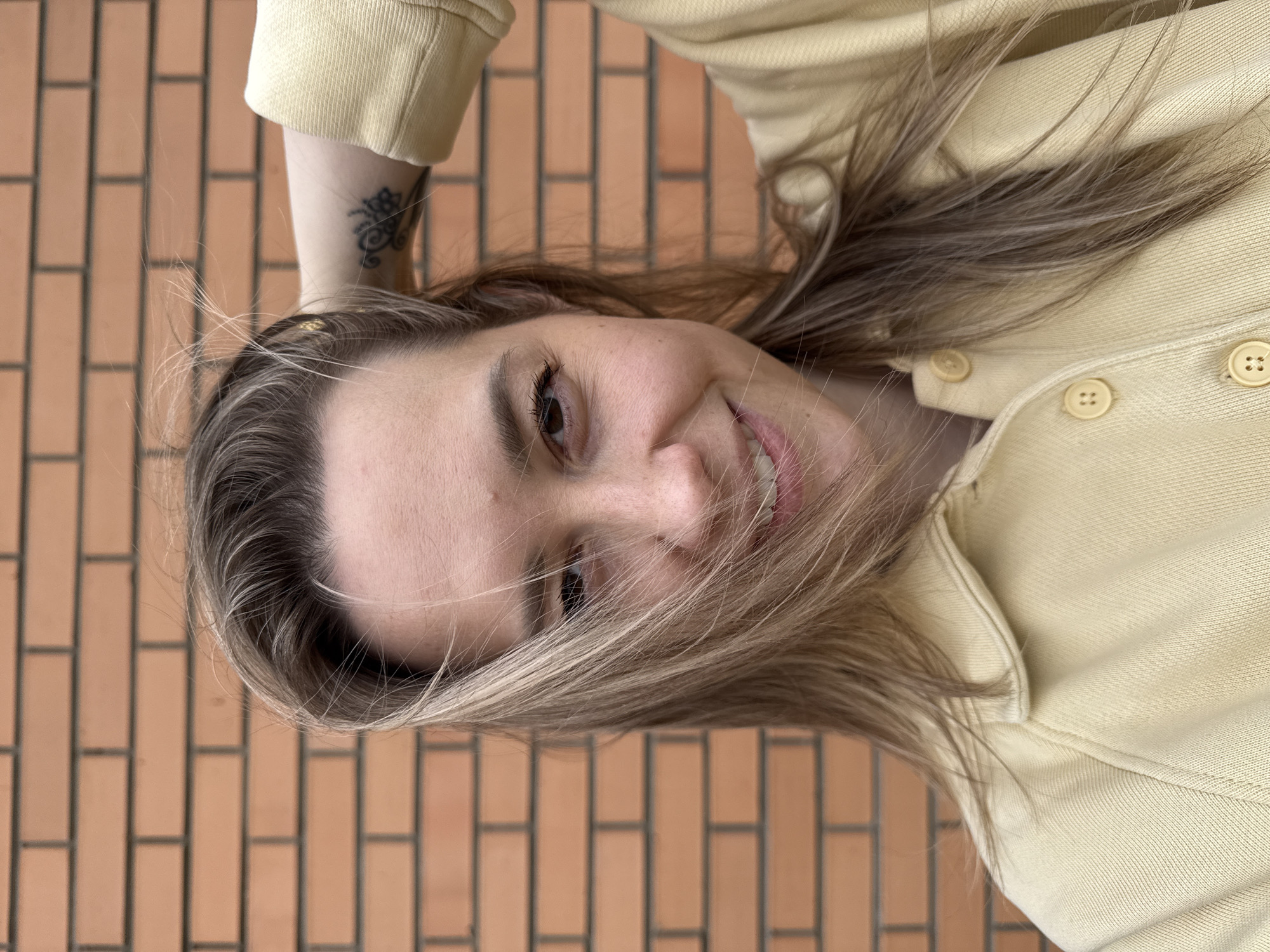 An image captures a young woman from the chest up, looking directly at the camera with a warm smile. Her head is tilted slightly, and her long, multi-toned blonde and light brown hair, with visible darker roots, falls casually around her face, with some strands gently obscuring parts of her forehead and cheek. She appears to be in her 20s or early 30s.
She is wearing a light yellow, textured button-up top or jacket, with several round, pale yellow buttons visible along the front placket. On her right arm, just below the sleeve of her top, a small, intricate dark-ink tattoo with an ornamental or floral design is visible.
The background consists of a uniformly patterned brick wall, composed of rectangular, reddish-brown bricks laid in horizontal courses with contrasting dark grout lines. This suggests an outdoor setting, possibly an exterior wall of a building. The lighting is bright and natural, indicating it is daytime. The woman's expression is friendly and engaging, conveying a positive emotion. There is no visible text in the image. The scene is located in Chaikovsky, Russia.