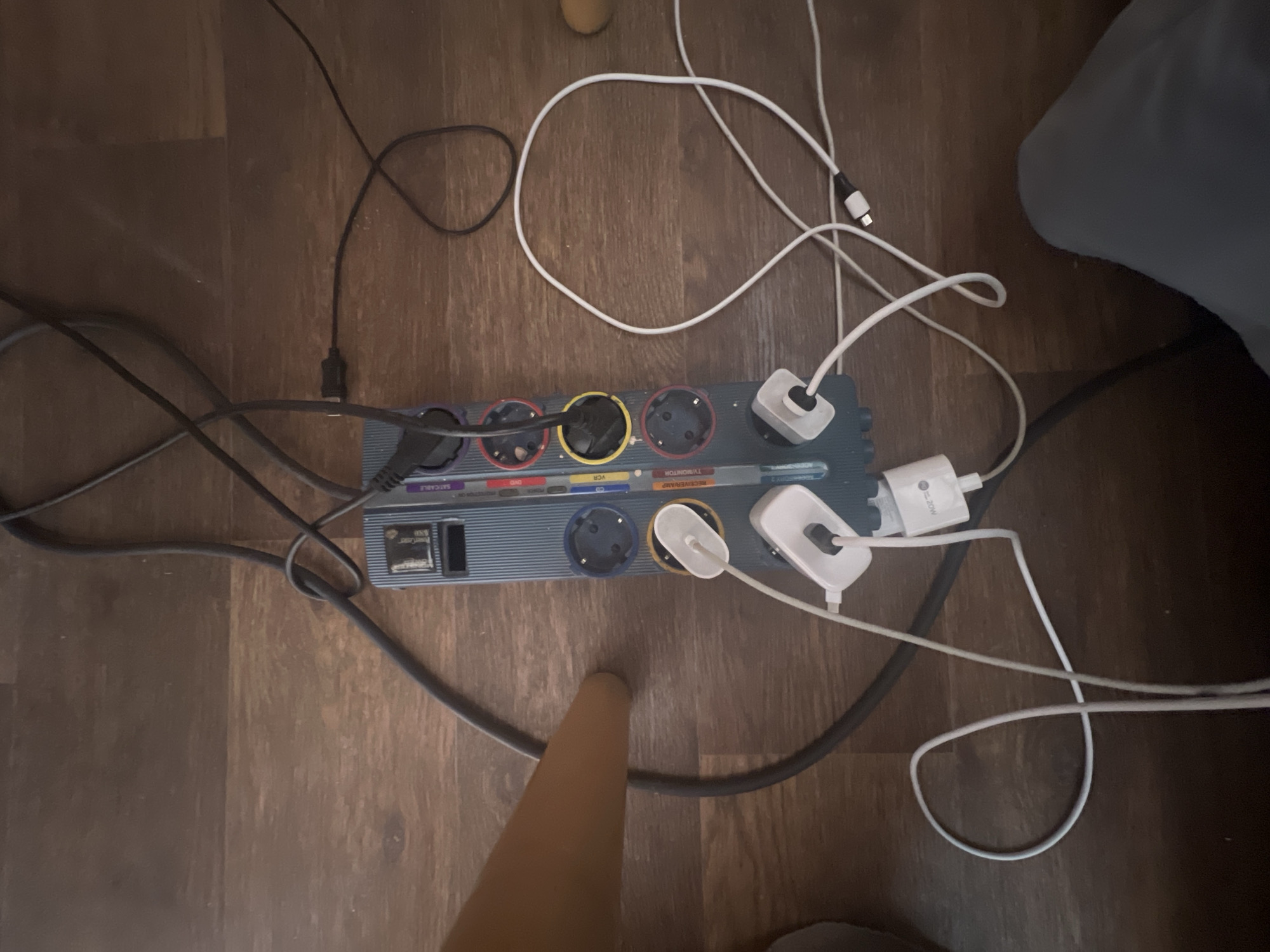 The image shows a power strip with multiple plugs and cables on a wooden floor. The power strip is blue and has labels for various devices like "DVD," "VCR," "TV/MONITOR," and "RECEIVER/AMP." Several white and black cables are plugged into the power strip, some connected to white charging bricks. A black cable from the power strip snakes across the floor and loops towards the top of the image. In the foreground, a person's legs, clad in gray fabric, are visible. A portion of a light brown wooden chair leg is on the left side of the image. The lighting suggests an indoor environment. The scene depicts a common household or office setup with electronic devices connected to power. There is no information in the image to suggest the location in the city of Трещиха, Russia.