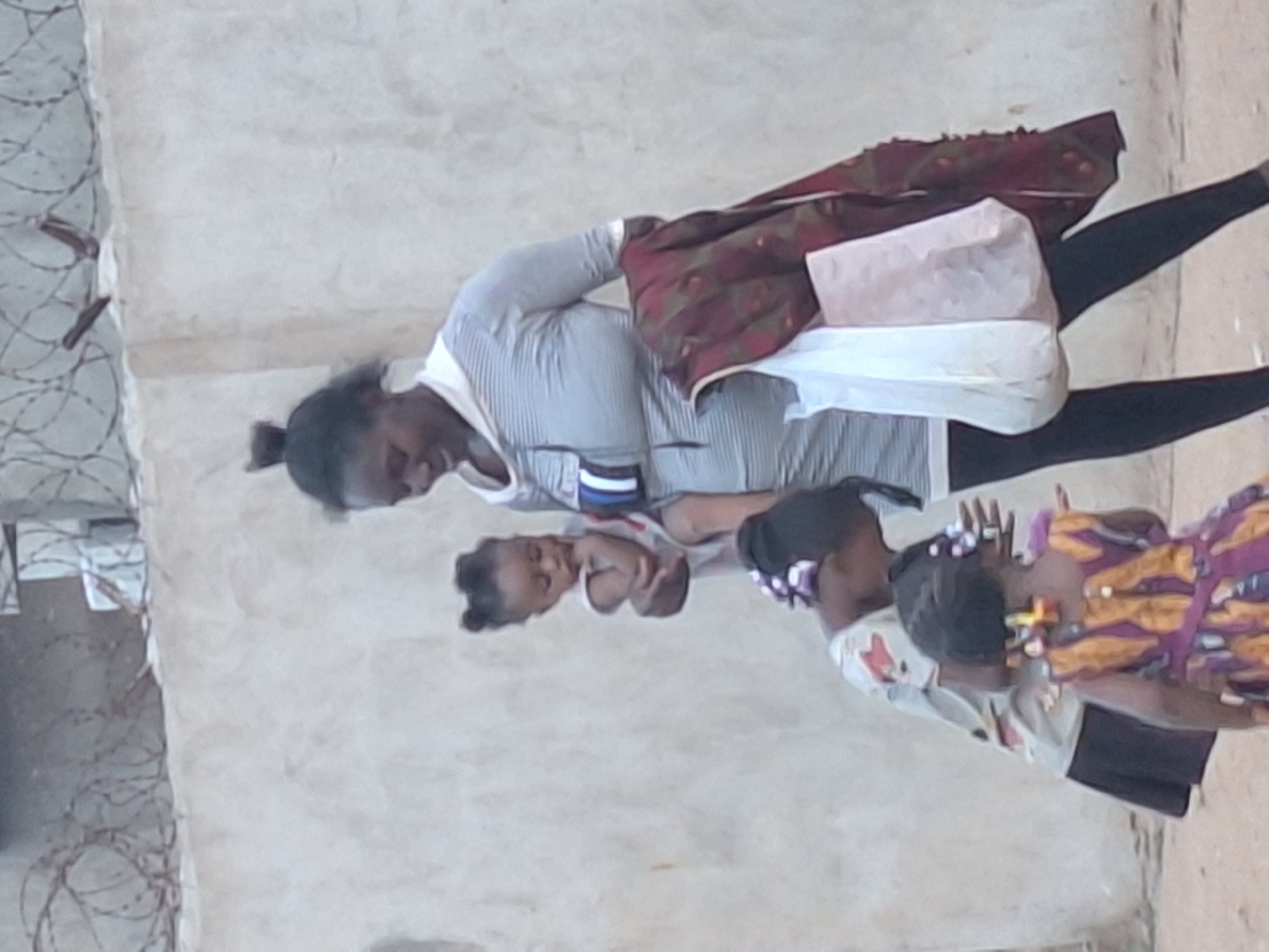 An adult woman holding a baby is shown standing with two young girls in an outdoor setting in Sabon Tasha, Nigeria. The image is taken from a slightly elevated perspective.
The woman, who appears to be of African descent, has dark hair tied back and is wearing a light grey and white striped long-sleeved top and dark pants. A patterned, possibly reddish-brown cloth with a decorative motif is draped over her left shoulder. She carries two light-colored bags in her left hand, one appearing to be a paper bag and the other a white plastic bag. She is holding an infant, who is wearing a white top, in her right arm. The baby appears calm.
Beside them are two young girls, also of African descent. One girl wears a white t-shirt with a small graphic design and dark lower garments (shorts or a skirt). The other girl is dressed in a vibrant patterned dress featuring shades of purple, yellow, and brown. Both girls have dark hair, with some accessories or beads visible in their hairstyles. Their faces are partially obscured or turned away, but they seem to be observing their surroundings or the woman and baby.
The background consists of a light-colored, possibly concrete, wall or building surface. On the far left, a section of wall is topped with barbed wire, suggesting an enclosed area or security measure. The ground appears to be an unpaved or dirt surface. The lighting suggests it is daytime, but it is somewhat muted, possibly indicating an overcast sky or shaded area. No discernible text is visible on clothing or in the environment. The overall mood is casual and observational, depicting an everyday scene of a family group together.