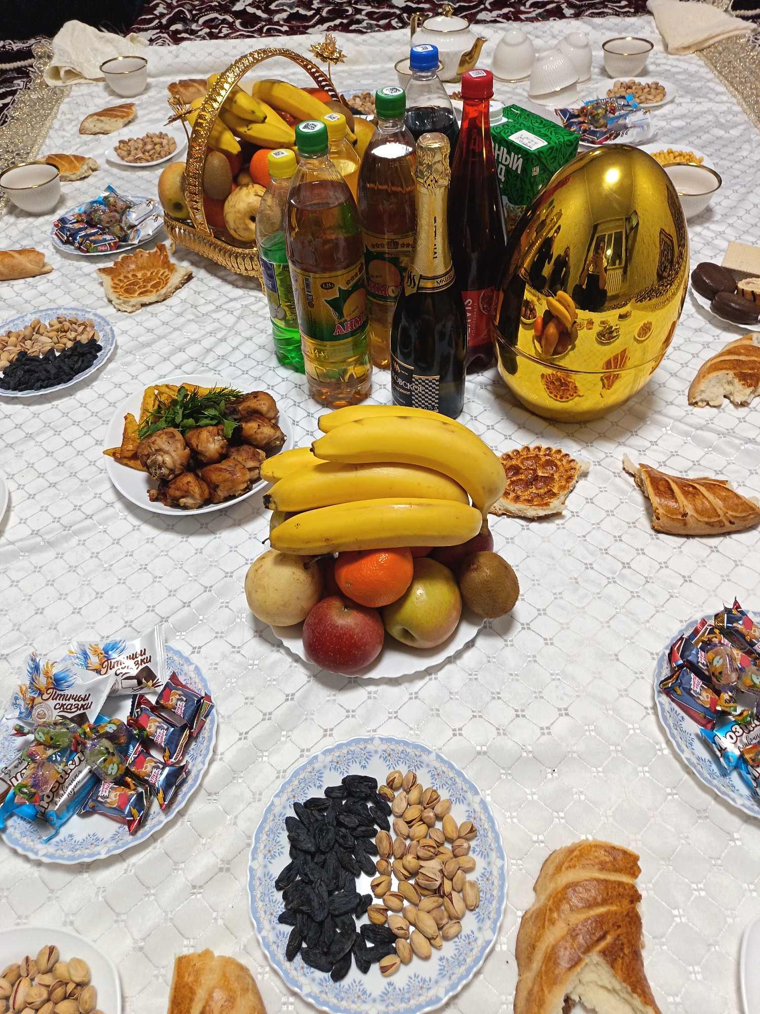 The image shows a spread of food and drinks on a white tablecloth. In the center, there is a basket filled with bananas, oranges, apples, and kiwis, surrounded by various beverages including bottled juices, sodas, and a bottle of sparkling wine. A large golden egg-shaped container reflects the surroundings.
The setting includes plates of assorted treats such as dried fruits, nuts, candies, pastries, and pieces of bread. On one plate, there are fried chicken pieces garnished with herbs. There are also teacups and a teapot.
Based on the beverage labels and candy wrappers, the location may be in Russia. However, no features specifically indicate Nizhnevartovsk.