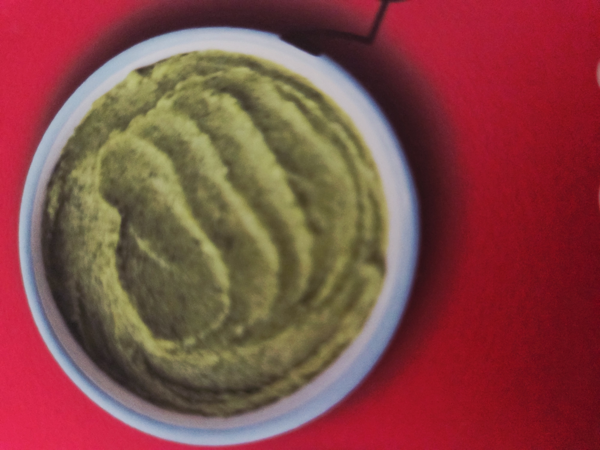 The image depicts a bowl of green substance, possibly mashed avocado or a similar food, set against a red background. The bowl appears to be white with a light blue rim. The substance inside has a ridged or swirled pattern. The photograph is taken from a slightly high angle. There is a small dark shape in the top-right corner. There is no information to suggest this image was taken in Kyustendil, Bulgaria.