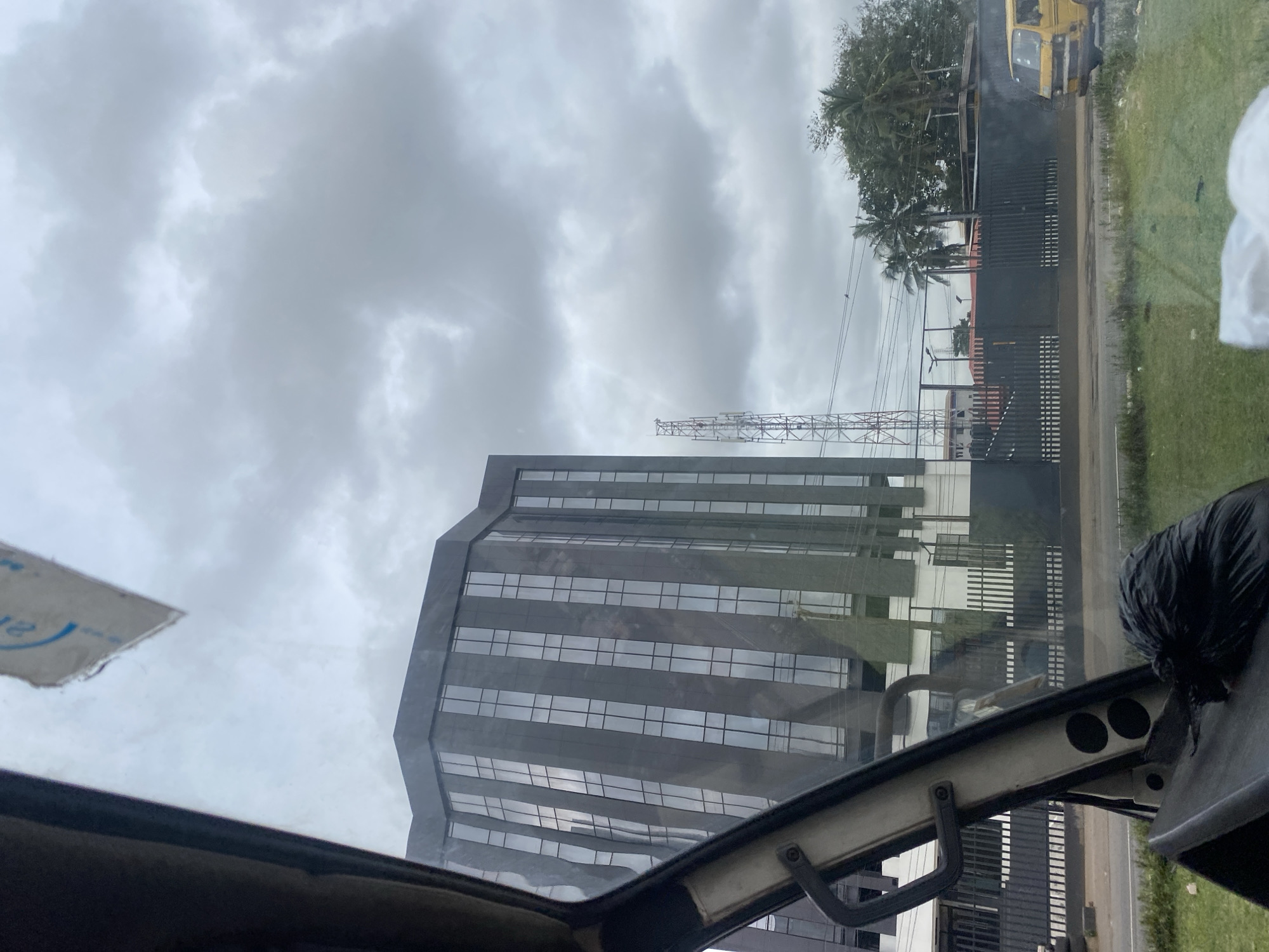 The image captures a view from inside a vehicle, looking out at an overcast urban environment in Ikeja, Nigeria. A modern, multi-story building with a dark, reflective facade and prominent vertical window frames dominates the center-left of the frame. To its right, a tall, lattice-like communication tower rises against the cloudy sky. A dark, imposing fence runs across the middle ground, separating the building and tower from a street and a patch of grass in the foreground. A yellow minivan is parked on the right side of the street, partially obscured by the fence. The interior of the vehicle is visible in the foreground, with a black trash bag resting on a surface, and the edge of the windshield and dashboard creating a frame. The overall impression is one of a functional, albeit somewhat dreary, city scene under a cloudy sky.