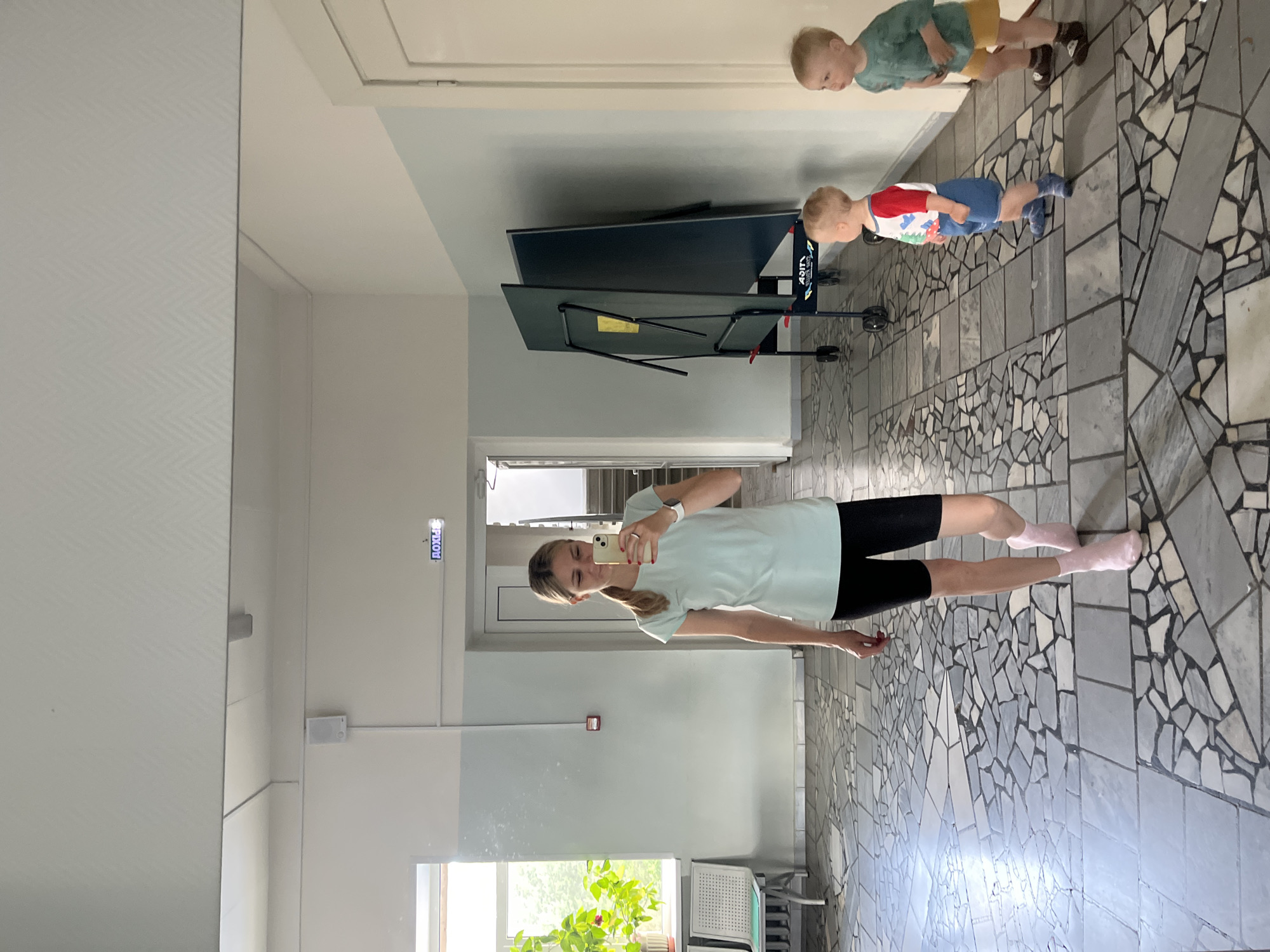 The image shows a woman taking a selfie in a long hallway. She is wearing a light blue t-shirt, black shorts, and pink socks. She has her hair pulled back in a ponytail and is wearing a watch on her left wrist. In the background, there are two young boys. One boy is wearing a green t-shirt and yellow shorts, and the other is wearing a red and white t-shirt and blue shorts. They appear to be standing near a folded table tennis table. The hallway has a tiled floor with a mosaic-like pattern of grey and white tiles. There are light blue and white walls, and a large window is visible on the left side of the image, with a plant visible through it. An exit sign with Russian text reading "ВЫХОД" (VYKHOD, meaning EXIT) is visible on the wall. The lighting suggests it is daytime. The overall setting appears to be an indoor common area, possibly in a school or community center. The woman's expression is neutral. The scene is captured in portrait orientation, rotated 90 degrees counter-clockwise.