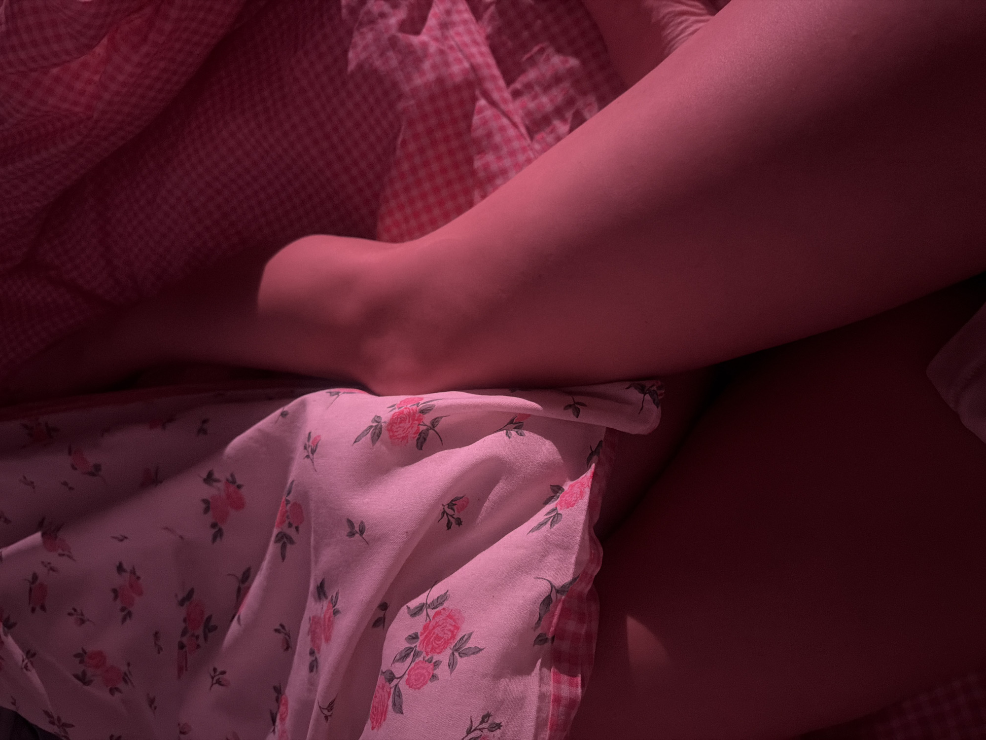 A close-up shot focuses on a person's leg and ankle, partially covered by bedding. The bedding includes a pink and white gingham patterned blanket and a floral patterned sheet with pink roses and green leaves. The lighting is subdued, casting a pink hue over the scene. There is no indication of the city of Moscow.