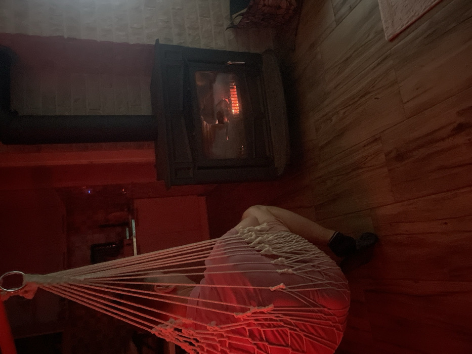 An indoor scene in Palež, Montenegro, displays a seated individual in a white macrame-style hanging chair, viewed from behind. The person wears a light-colored upper garment and dark socks, facing a dark-toned, corner-positioned wood-burning stove with visible flames behind its glass door. The room's floor is composed of wooden planks, and a segment of a white brick wall is visible behind the stove. The ambient lighting throughout the space exhibits a prominent reddish hue. In the distant background, a dark object resembling a screen is faintly discernible. A dark woven basket and a light-colored rectangular rug are partially visible on the floor to the right of the stove.