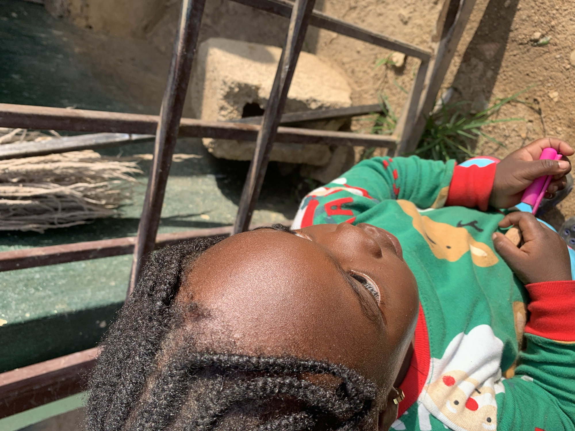 A young child, with dark skin and braided black hair, is captured in a close-up, high-angle shot, lying down outdoors. The child is wearing a vibrant green long-sleeved pajama top featuring Christmas-themed motifs, including a prominent brown reindeer and a white Santa Claus. The cuffs of the pajamas are red with green and white stripes. In the child's right hand, a pink and light blue plastic object, possibly a toy, is held.
The child's eyes are partially closed or gazing downwards, with visible eyelashes. The forehead and skin appear smooth and well-lit by bright sunshine. The posture suggests a moment of rest or quiet observation.
The setting is an outdoor area, indicating a clear, sunny day. The background features rusty metal bars, likely part of a fence or gate, running across the frame. Below these bars, a bundle of dry, straw-like material is visible. Further back, concrete blocks are scattered on dry, light brown earth, interspersed with sparse patches of green grass. The scene evokes an open, uncultivated environment in New Stadium, Nigeria. There is no visible text in the image, and the child's expression is calm.