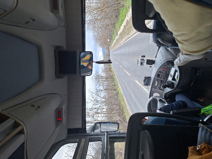 The image depicts the interior perspective from a moving bus or coach in Gabarevo, Bulgaria, looking forward through the windshield onto a two-lane asphalt road. The road is lined on both sides by leafless deciduous trees and some green undergrowth, indicative of late autumn, winter, or early spring. An overcast sky is visible above.
In the foreground, the lower legs and feet of a passenger, wearing light-colored pants and beige shoes, are elevated and resting on the right side of the vehicle's interior, near the dashboard. To the left, the bus driver is partially visible, wearing a blue long-sleeved shirt and blue jeans, with hands on the steering wheel, actively operating the vehicle. The dashboard features a steering wheel, a gear selector, instrument clusters, various buttons, a mounted GPS device, and an action camera.
Overhead, a small black monitor is centrally mounted, below which a rearview mirror reflects a yellow object and a partial human head. Storage compartments and a red illuminated indicator light are visible on the overhead panel. A green shopping bag and a brown textile item are situated on the floor or seat to the lower left. In the distance on the right side of the road, a circular red-and-white prohibitory road sign is present. Distant mountains are faintly visible under the overcast sky.