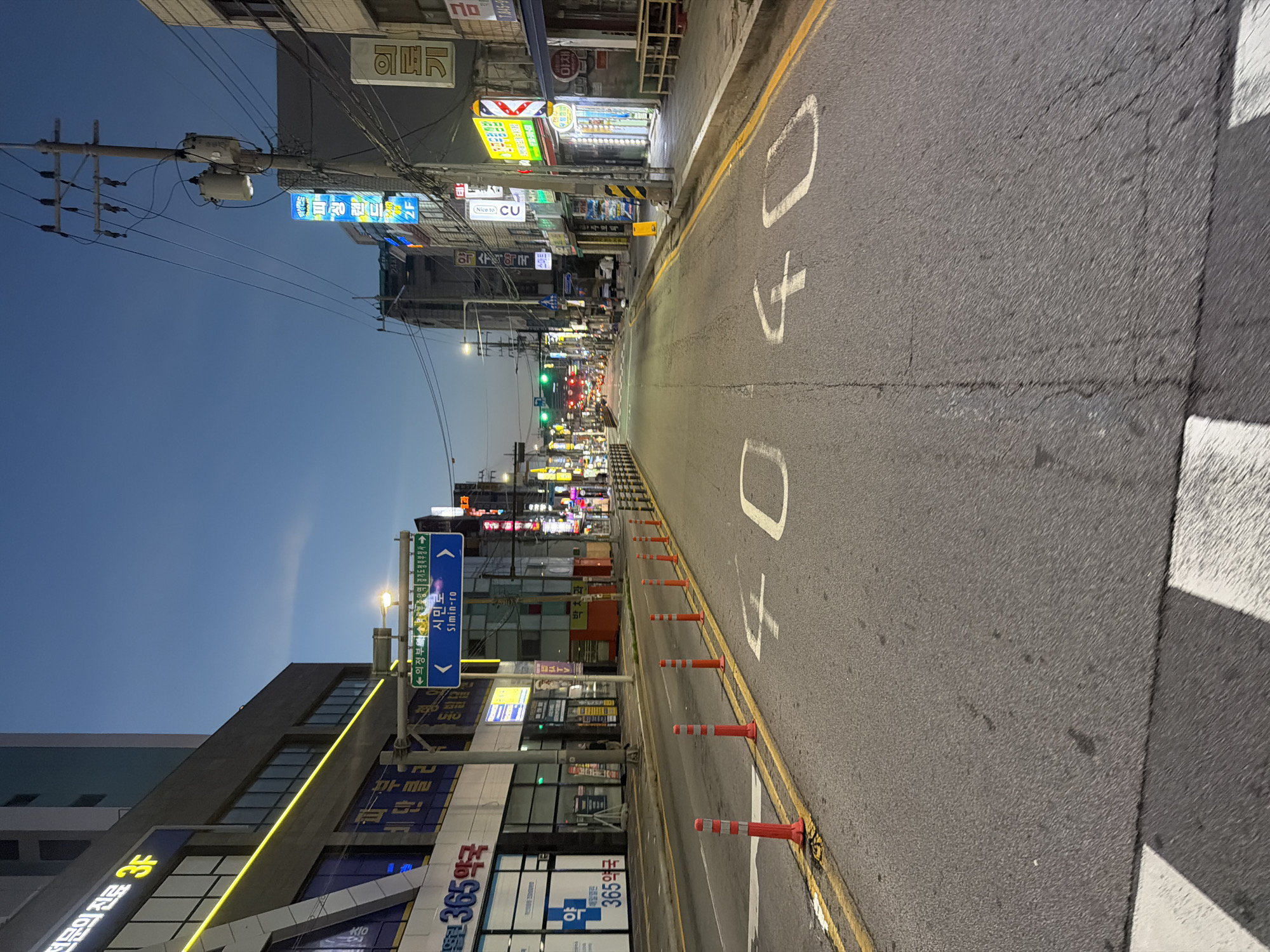 The image shows a street scene in Uijeongbu-si, South Korea, at dusk. The street is paved and has yellow lane markings. Traffic cones are placed along the right side of the road. White numbers "40" are painted on the road surface, indicating a speed limit. Buildings line both sides of the street, featuring various signs and illuminated windows. A traffic light is visible in the distance, showing a green light. The sky is a deep blue, suggesting twilight. There are no people clearly visible in the foreground.