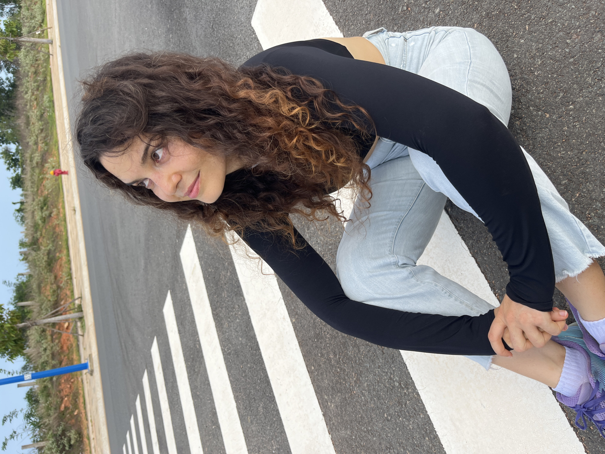 A young woman with curly brown hair sits on a pedestrian crossing in Mui Ne, Vietnam. She is wearing a black long-sleeved crop top and light-wash jeans. Her hands are clasped in her lap, and she is looking upwards with a slight smile. The pedestrian crossing is made of white stripes on an asphalt road. In the background, there is a sidewalk, a blue lamppost, some sparse vegetation, and a red fire hydrant. The sky is partly cloudy.