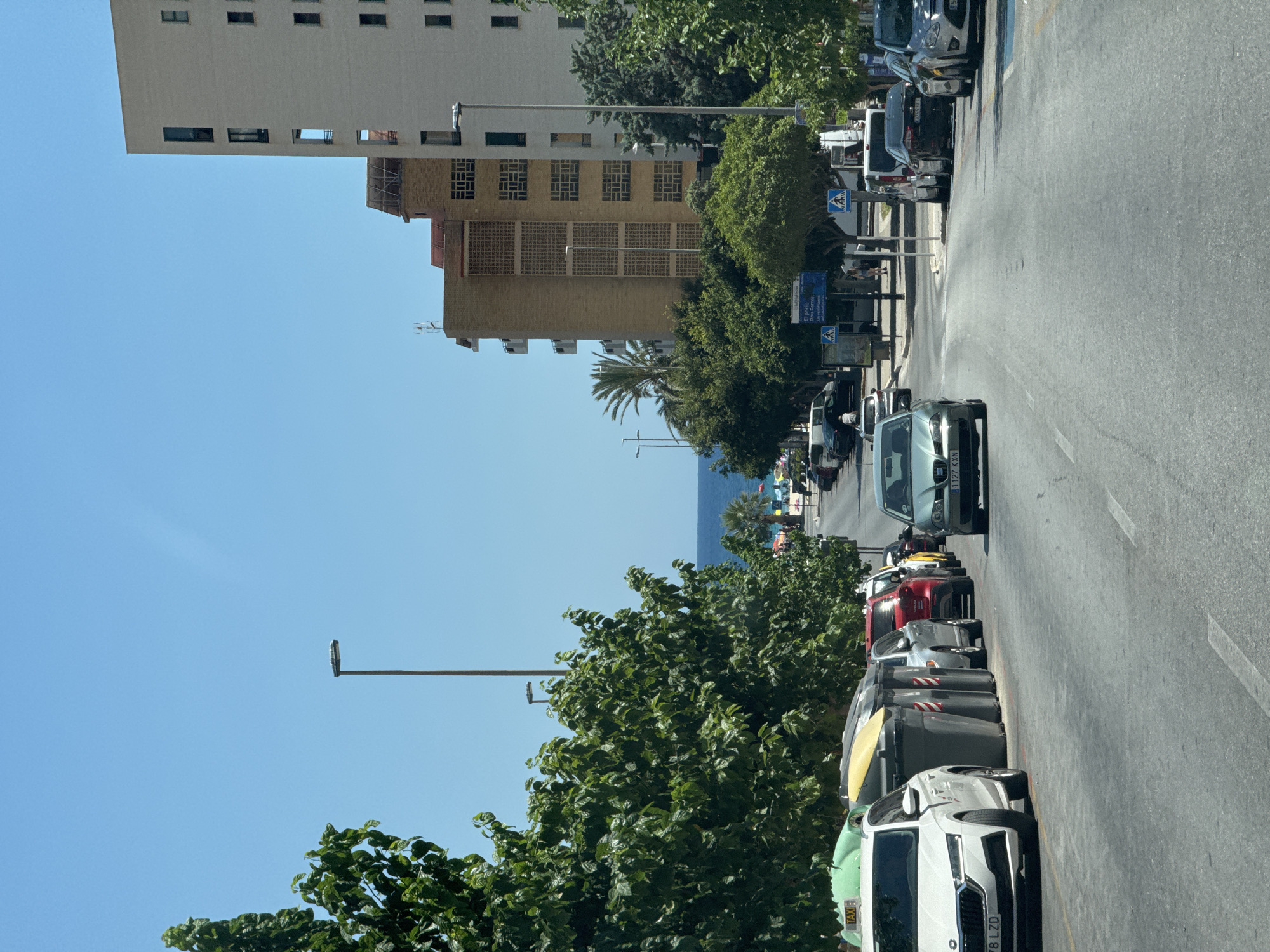 The image shows a street scene in La Vila Joiosa, Spain, on a clear, sunny day. The perspective is from a vehicle moving forward on an asphalt road. The street is lined with parked cars and large green recycling bins on the left, and several multi-story buildings and trees on the right. In the distance, the blue Mediterranean Sea is visible beyond a row of trees and some buildings.
On the left side of the street, a white Skoda sedan with license plate "78 LZD" is parked closest to the camera. Several large, grey recycling bins are lined up next to it. Further down the street, other cars are parked, including a silver Audi and a red car.
On the right side of the street, the buildings are modern, with visible balconies and windows. The closest building on the right is a light-colored, multi-story structure. Further down, there's a building with a distinctive yellow-brown facade and patterned window designs. Palm trees and other lush green trees line the sidewalk.
The road itself has white lane markings. There are pedestrian crossings indicated by blue signs with white walking figures. A few people are visible walking on the sidewalk in the distance. The sky is a clear, bright blue, with no clouds. The overall impression is a bright, urban coastal environment.