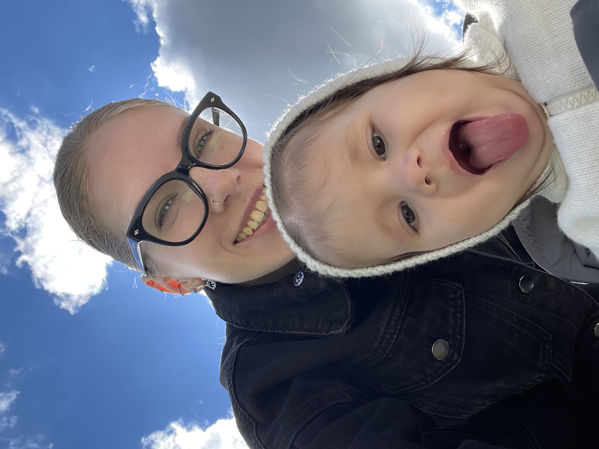 A low-angle selfie features an adult and a young child against a backdrop of a blue sky with white clouds. The adult, positioned on the left, is a fair-skinned woman with blonde hair pulled back. She wears black-framed eyeglasses, a nose ring, and multiple earrings in her left ear. The woman is smiling, revealing her teeth. She is wearing a dark denim jacket. To the right is a young child with brown hair and eyes. The child wears a white, knitted hat and a grey jacket and is sticking their tongue out. The photo's location cannot be determined to be Izhevsk, Russia, based on the visual information provided.