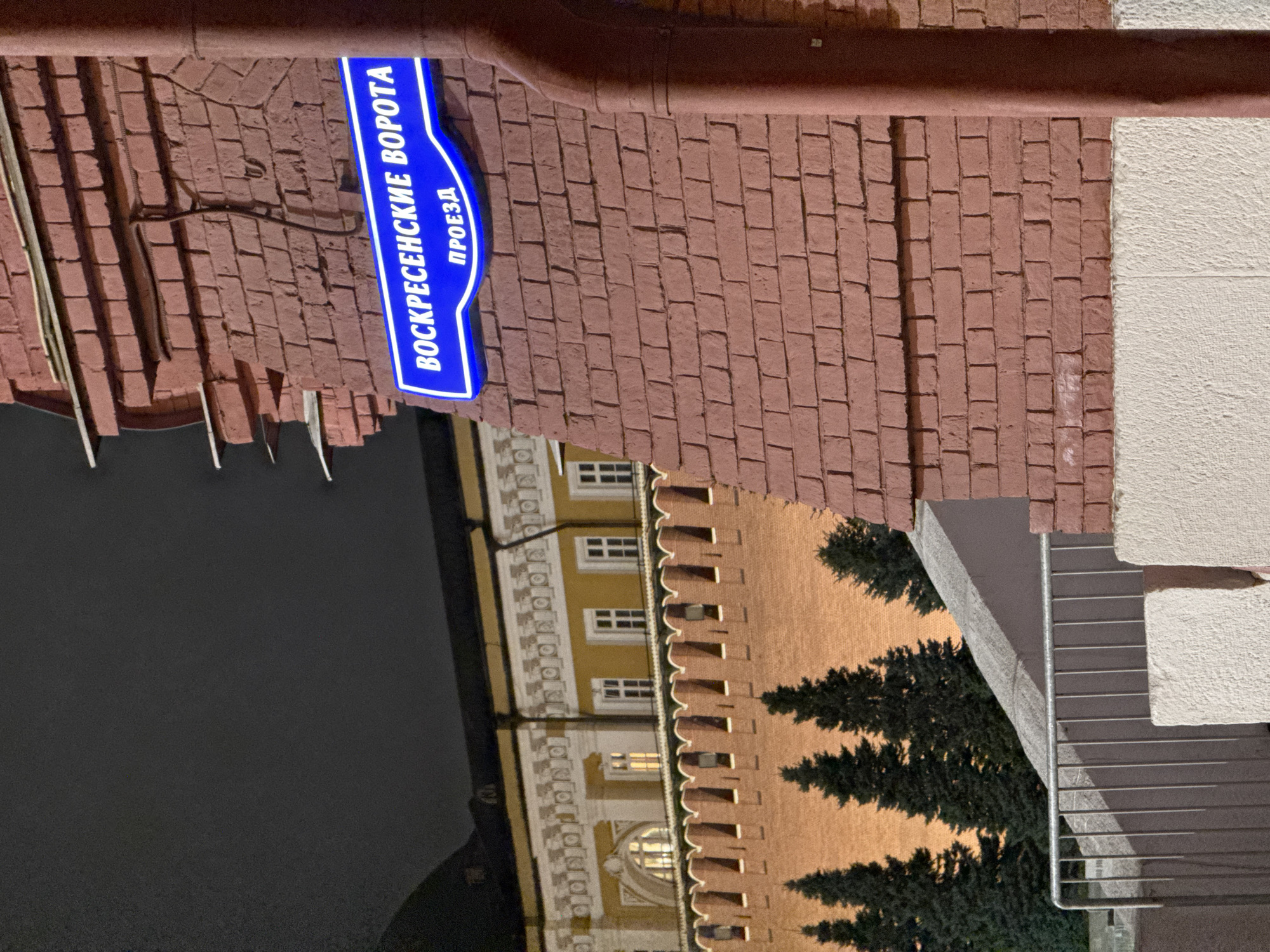 Here is a detailed description of the image:
The image captures a street sign that reads "Воскресенские Ворота Проезд" in blue lettering, affixed to a red brick wall. This indicates the location of Voskresensky Passage. In the background, a building with illuminated windows and architectural detailing is visible. A row of evergreen trees lines the base of the wall, adding a touch of nature to the urban setting. The image is slightly tilted, giving a unique perspective of the scene. The overall lighting suggests it's either late evening or nighttime.