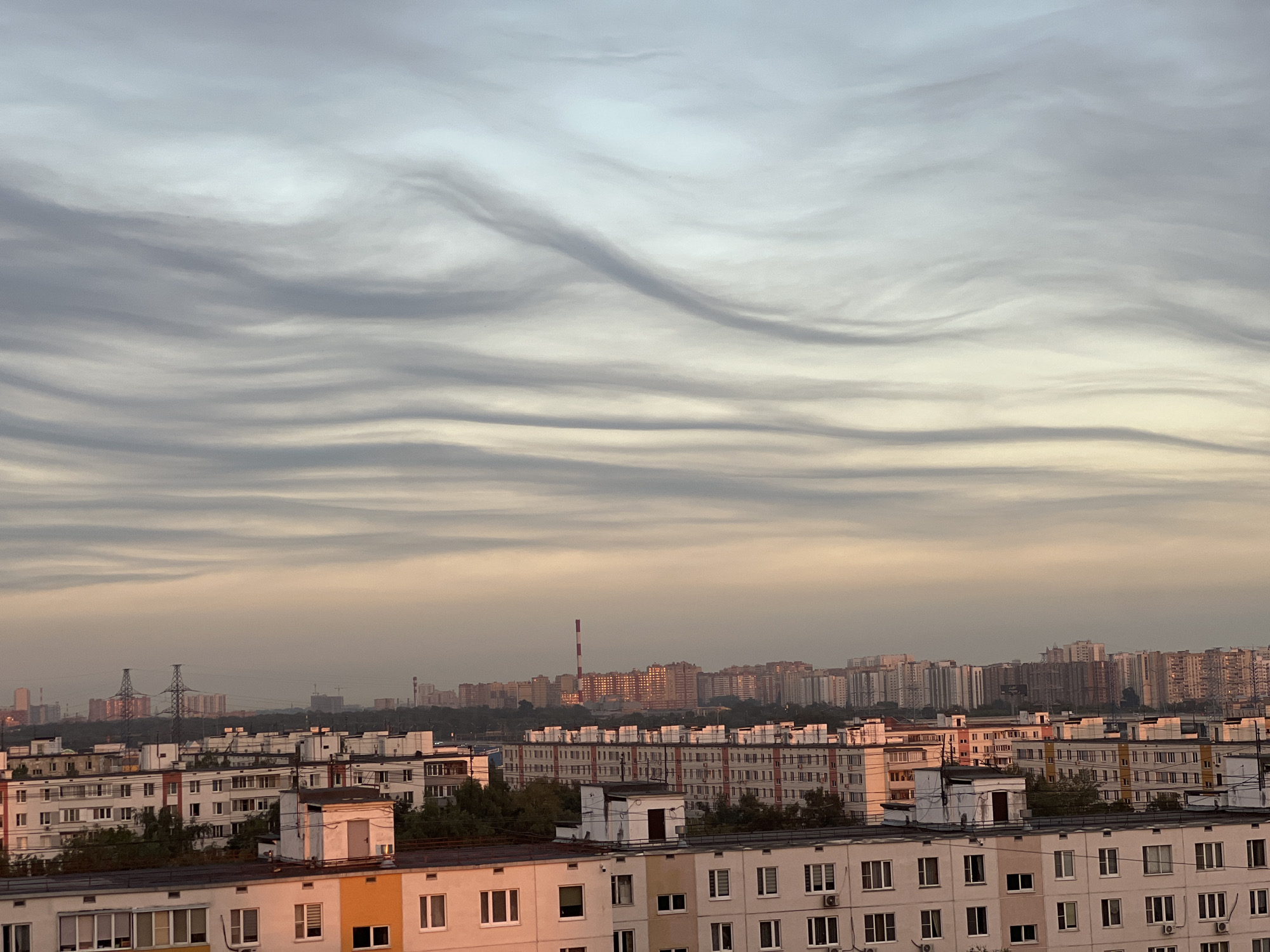 The image depicts a panoramic view of a densely populated urban area at dawn or dusk. The sky is filled with undulating, wave-like clouds that transition from pale blue at the top to a soft orange and pink hue near the horizon. Below the sky, a sprawling cityscape is visible, characterized by numerous apartment buildings of varying heights and architectural styles. Many of these buildings are constructed with concrete and feature repetitive window patterns. In the mid-ground and background, taller structures, including power transmission towers and industrial chimneys, are interspersed among the residential buildings, suggesting a mixed-use urban environment. The light conditions indicate either early morning or late evening, casting a warm glow on the facades of some buildings. There are no people or vehicles clearly visible in the frame. The scene appears to be a residential district within a large city, possibly Moscow, Russia, given the architectural style of the buildings and the presence of typical Soviet-era apartment blocks.